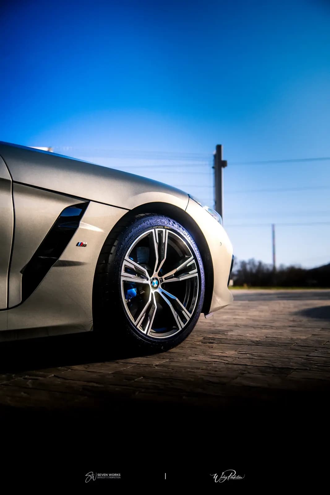 Detail předního kola BMW s modrou oblohou v pozadí, dramatické nasvícení.