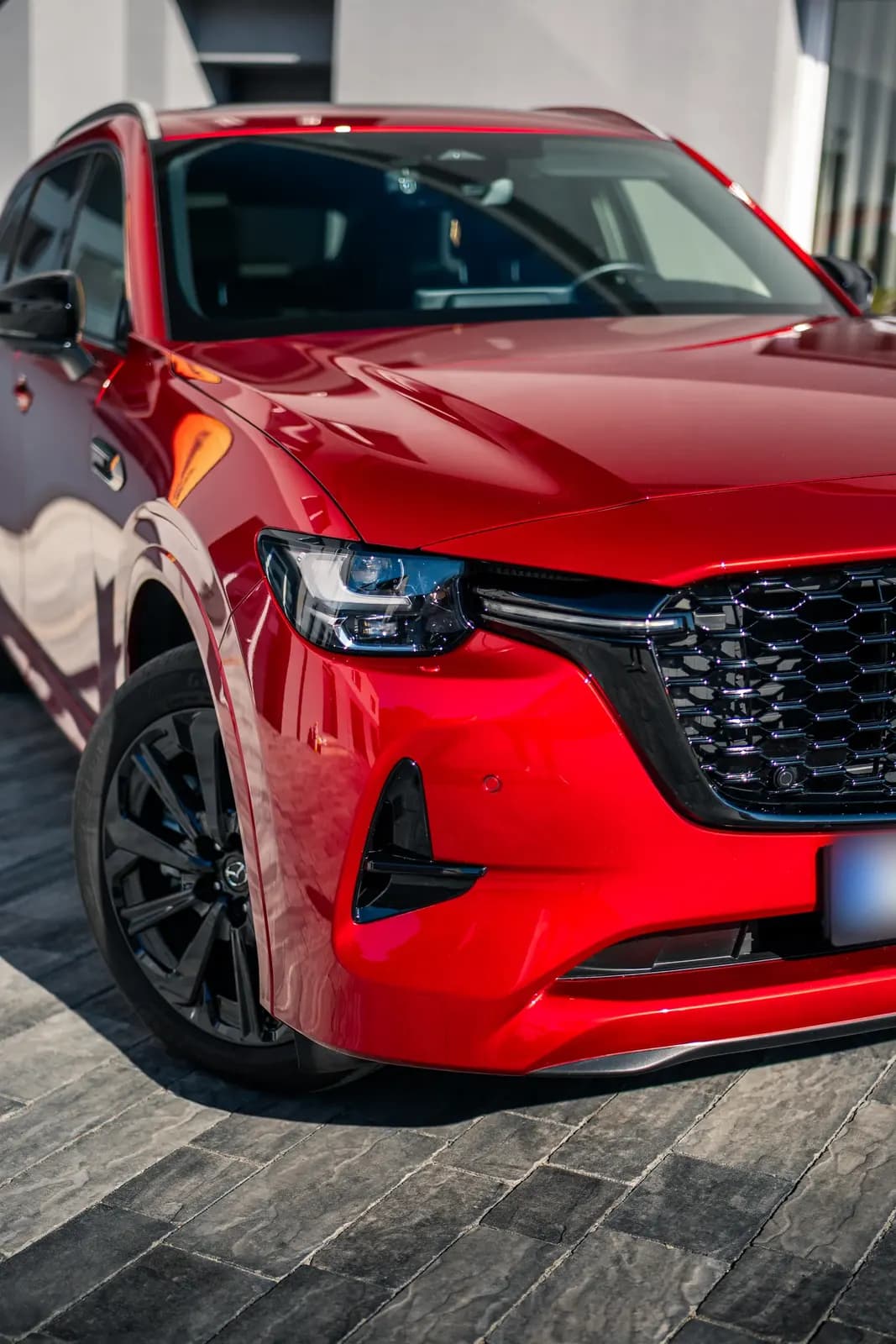 Detail předního nárazníku, světla a masky červené Mazdy CX-60 před budovou provozovny.