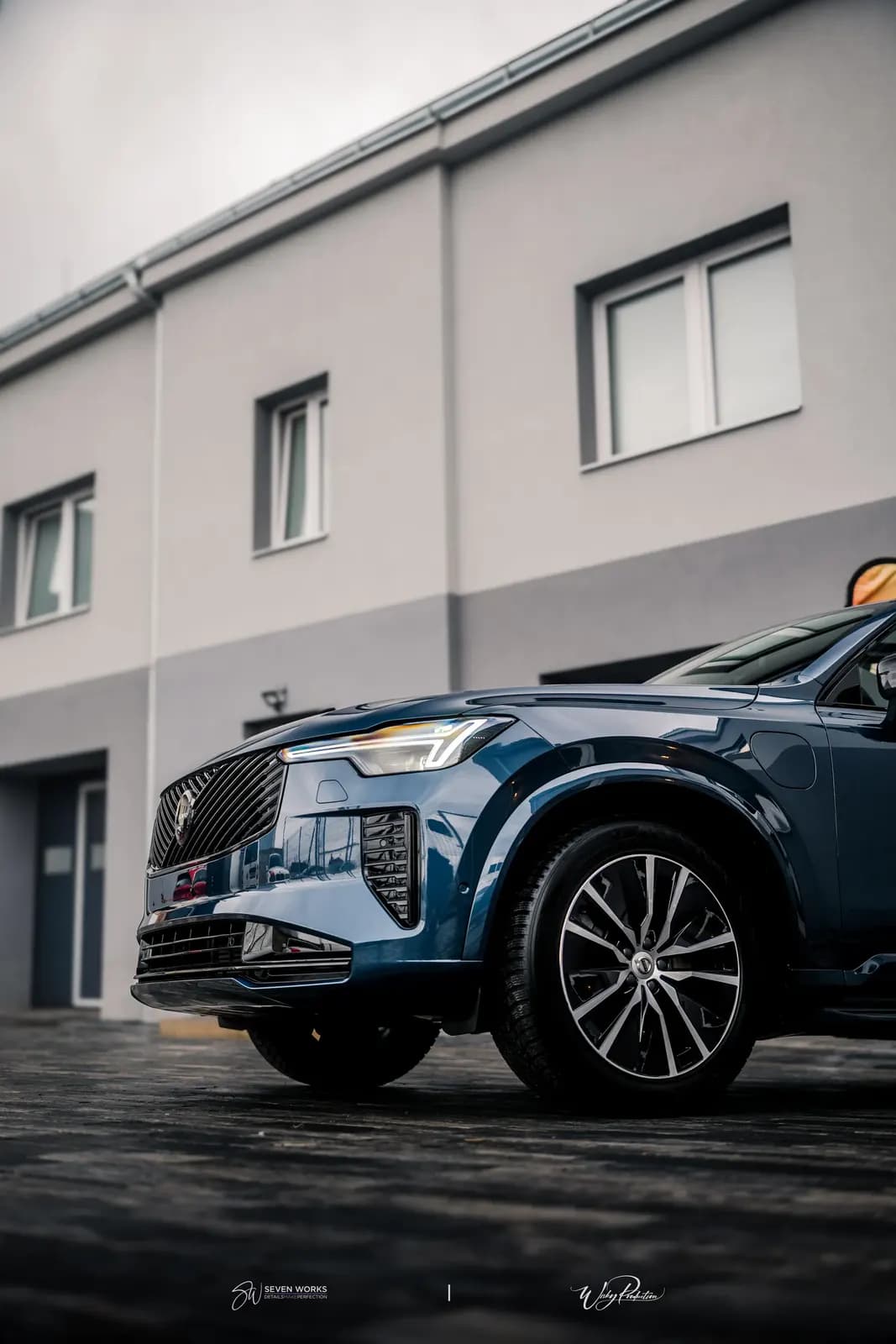 Modré Volvo XC90 zaparkované před provozovnou, záběr zespodu s oblačnou oblohou.