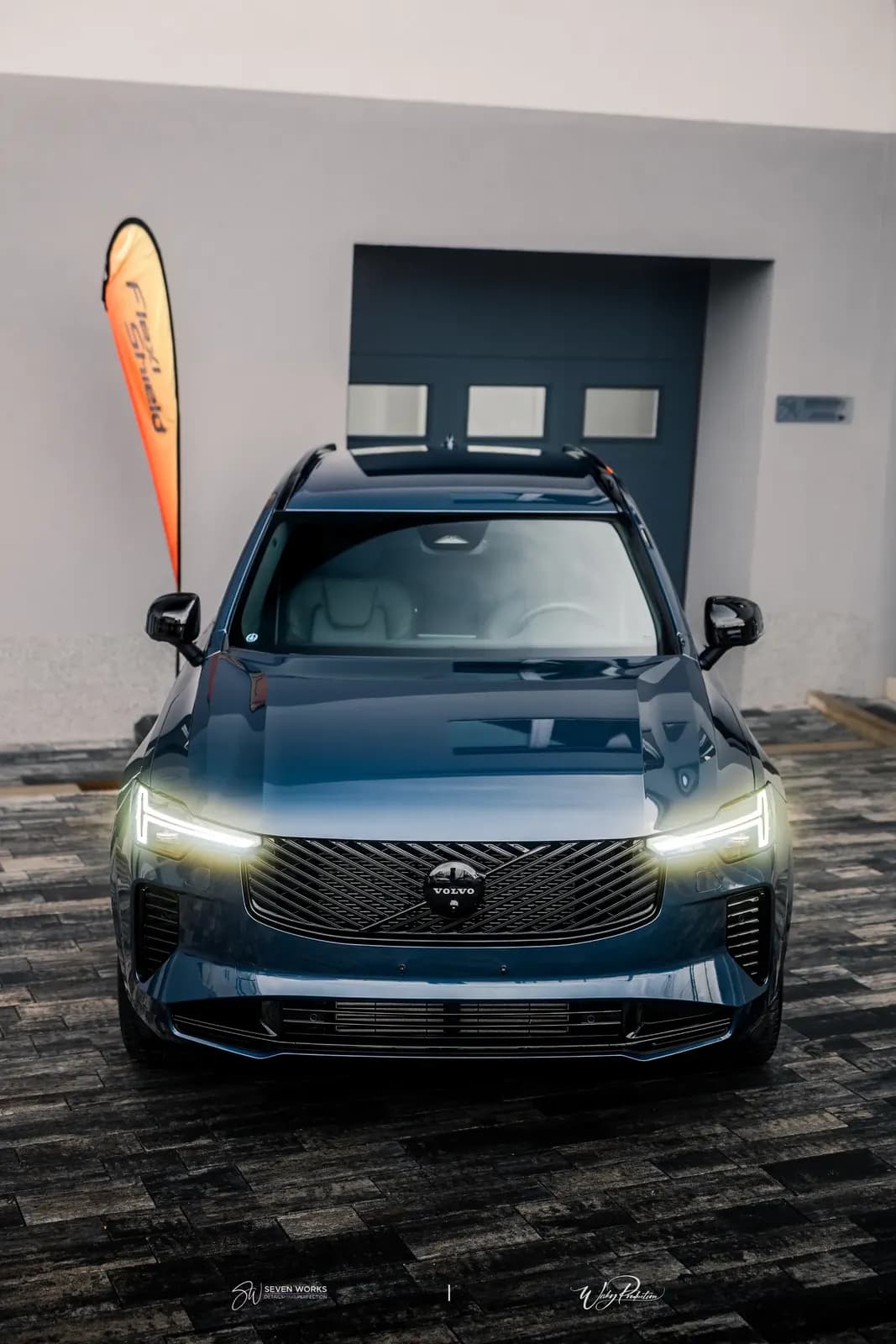 Volvo XC90 zepředu na nádvoří dílny, v pozadí garážová vrata a vlaječka FlexiShield.