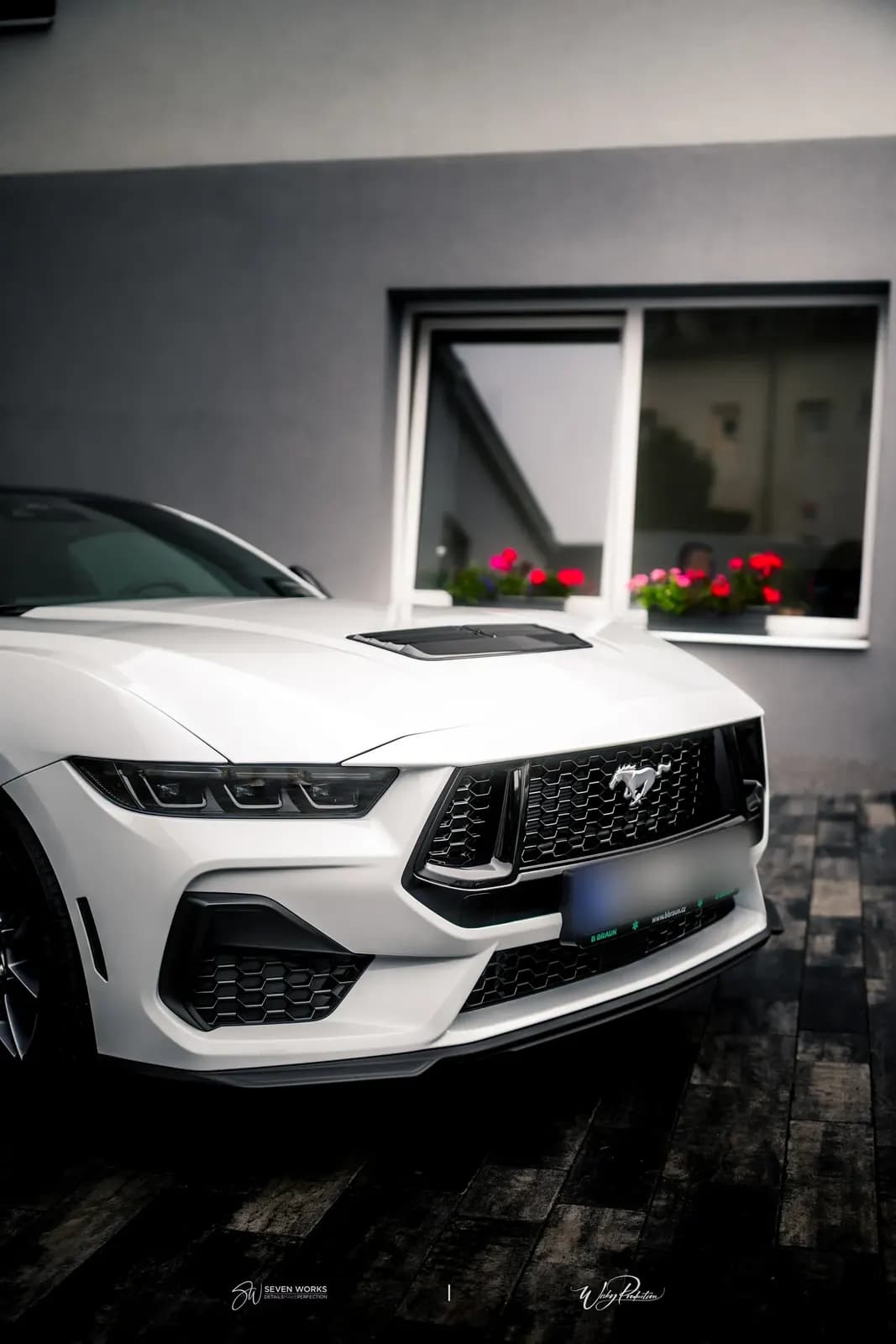 Detail přední kapoty a masky bílého Mustangu, v okně budovy za autem květiny.