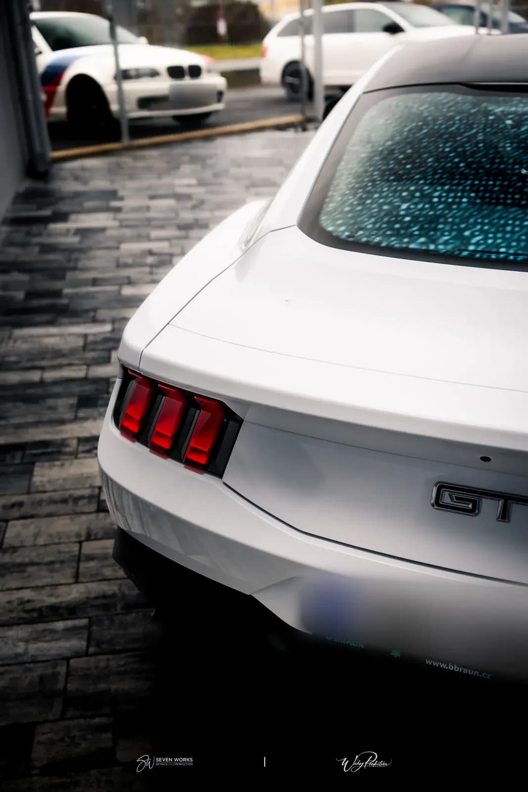 Detail zadních světel a nápisu GT na bílém Mustangu, v pozadí zaparkované vozy.