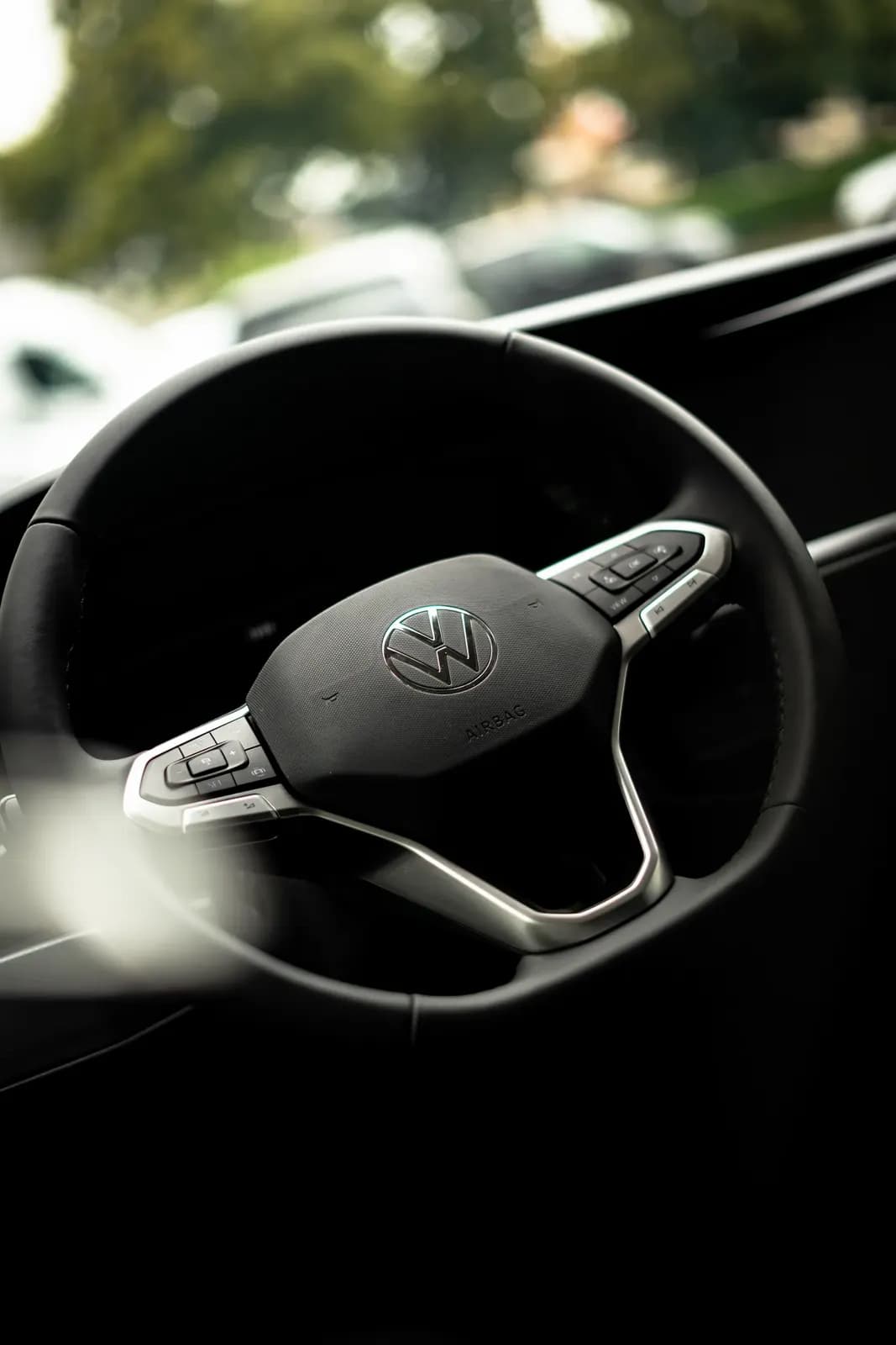 Volant Volkswagen s logem výrobce, v pozadí zeleň exteriéru.