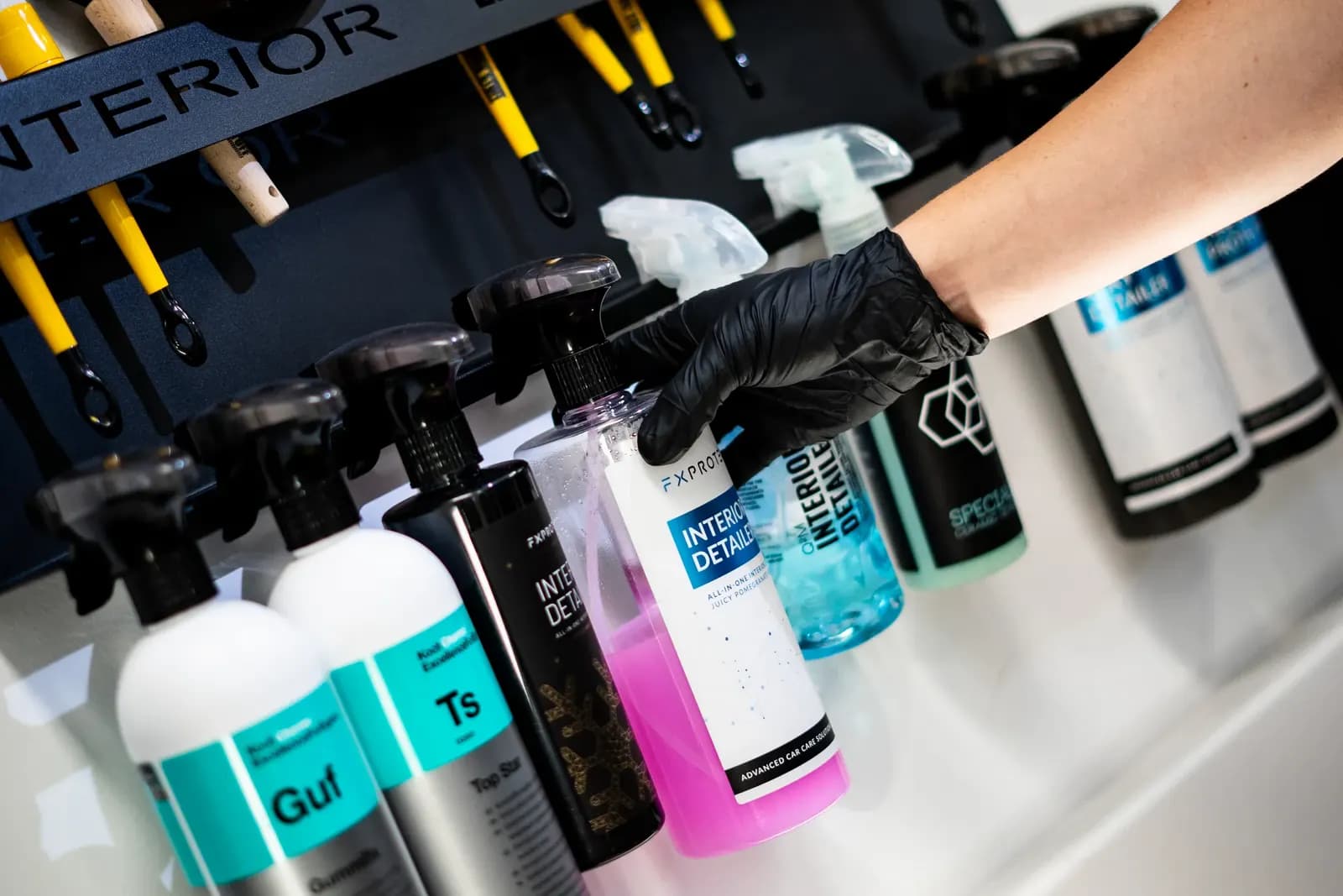 Ruka v rukavici sahá po lahvi čisticího přípravku na regálu s detailingovou chemií.