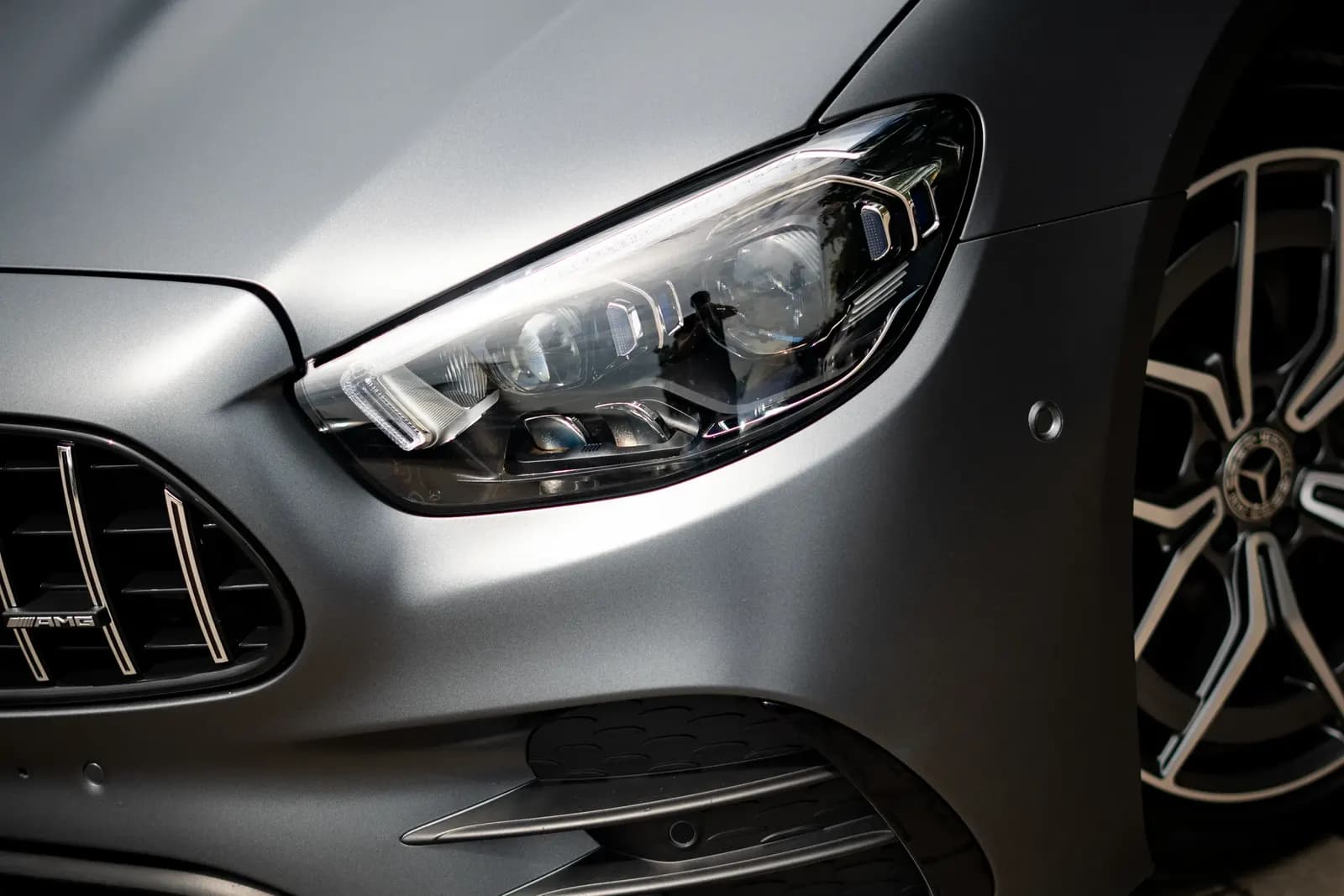 Close-up of the front headlight and grille with an AMG badge on a matte grey Mercedes sports car.