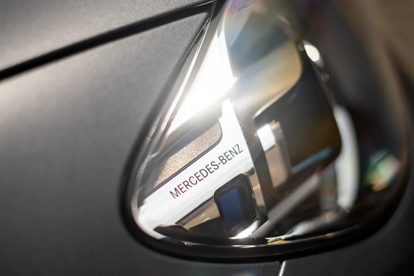 Close-up of a headlight with the Mercedes-Benz lettering on a matte grey car.