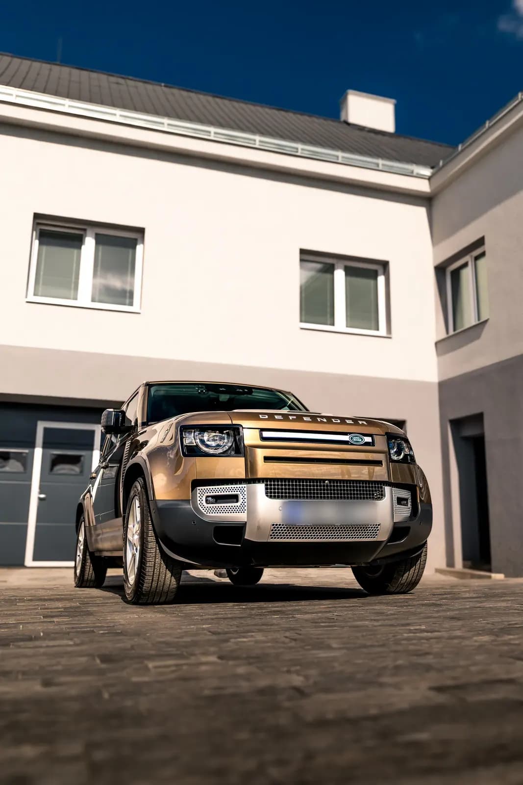 Land Rover Defender ve zlatohnědé barvě zaparkovaný před rodinným domem, pohled zepředu.