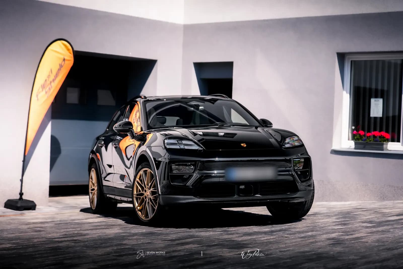 Black Porsche with bronze wheels in front of a garage, an orange flag on the left and a flower window on the right.