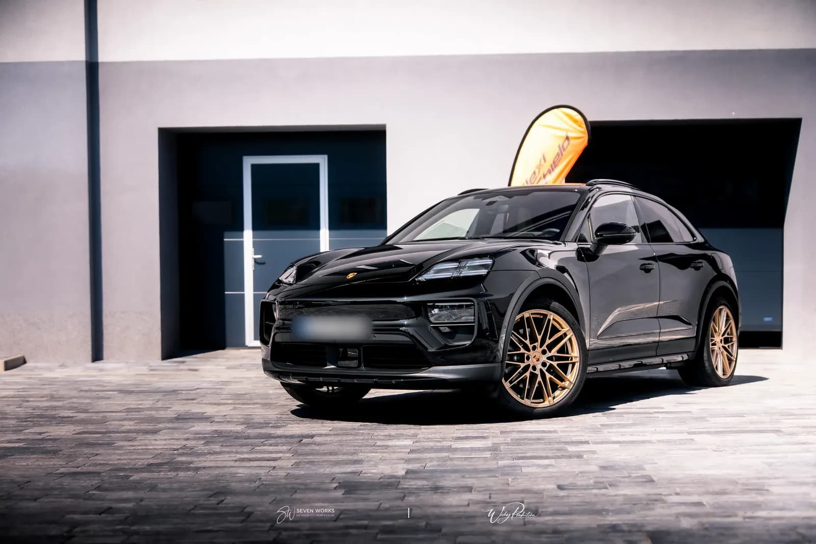 Front three-quarter view of a black Porsche with bronze wheels in front of a garage with an orange flag.