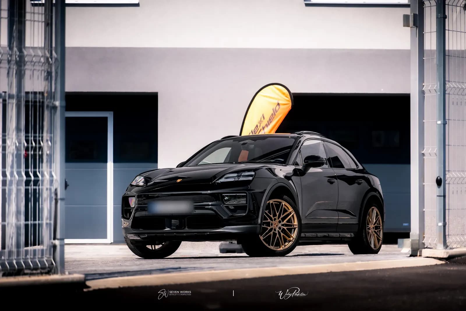 Black Porsche with bronze wheels parked behind a mesh gate, an orange flag behind it.