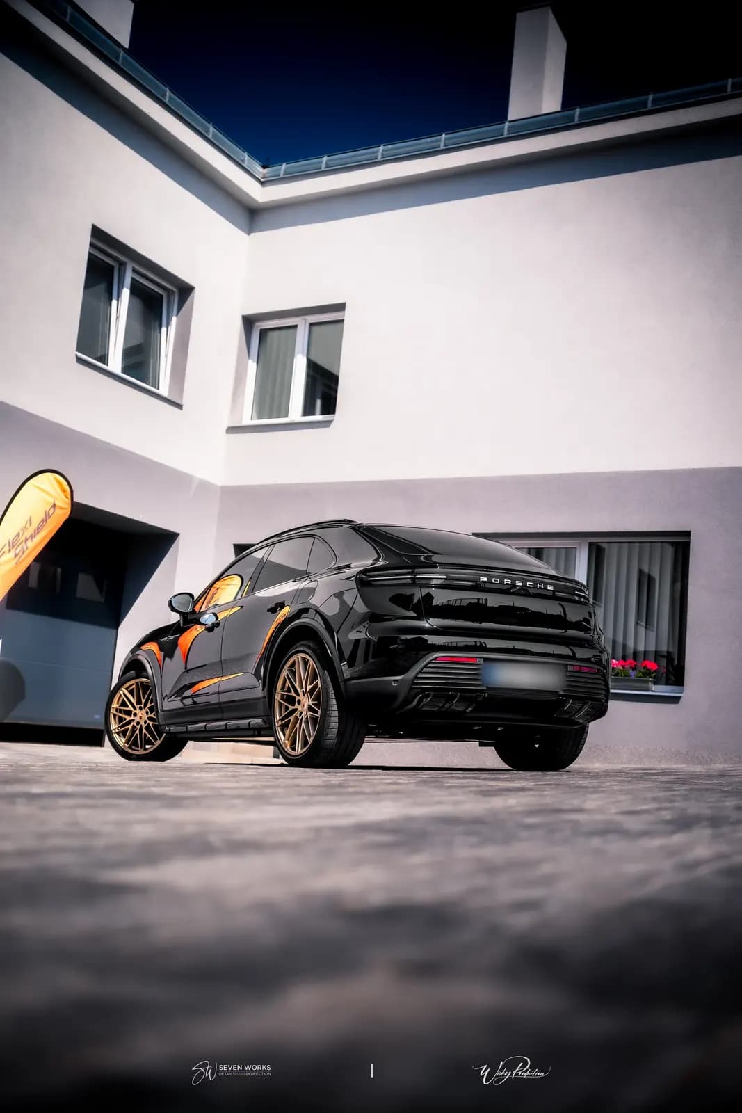 Rear three-quarter view of a black Porsche with the Porsche wordmark, a flower-filled window behind.