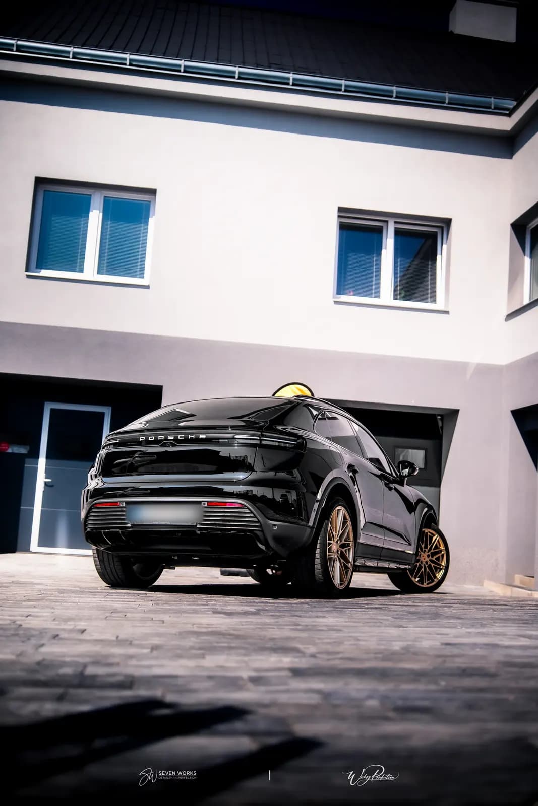 Rear view of a black Porsche with bronze wheels in front of a house facade with windows.