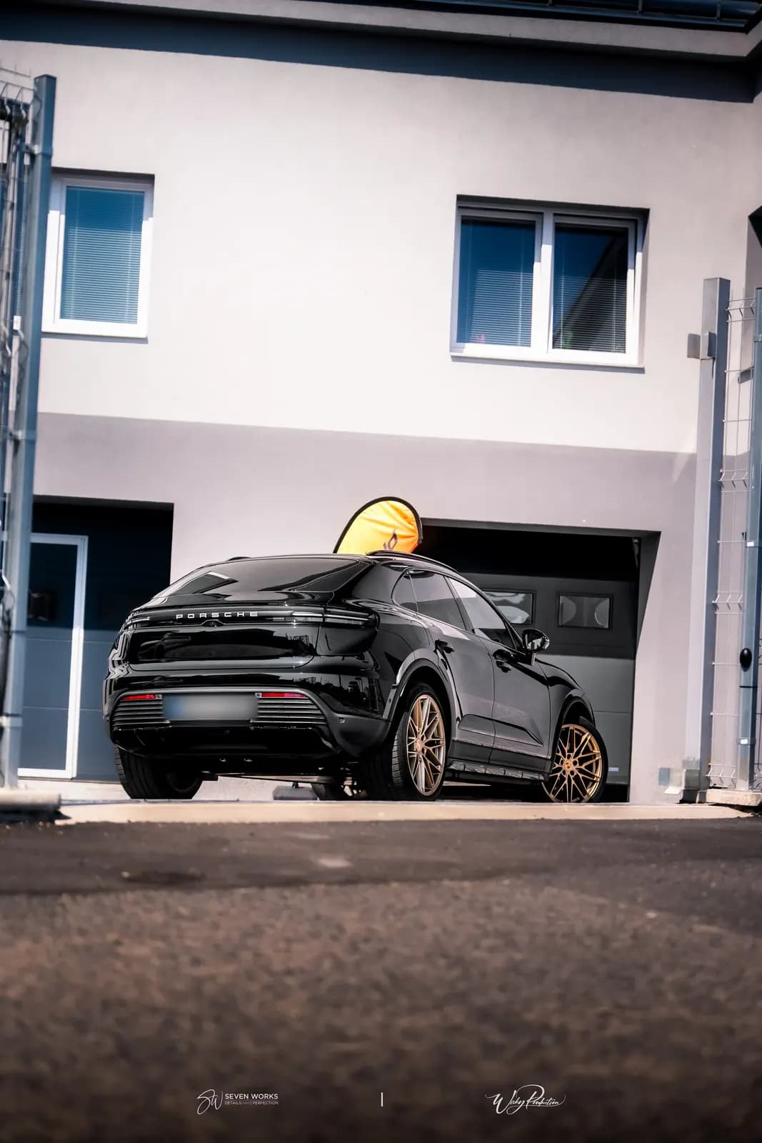 A black Porsche with bronze wheels behind a mesh gate, a yellow flag in the background.