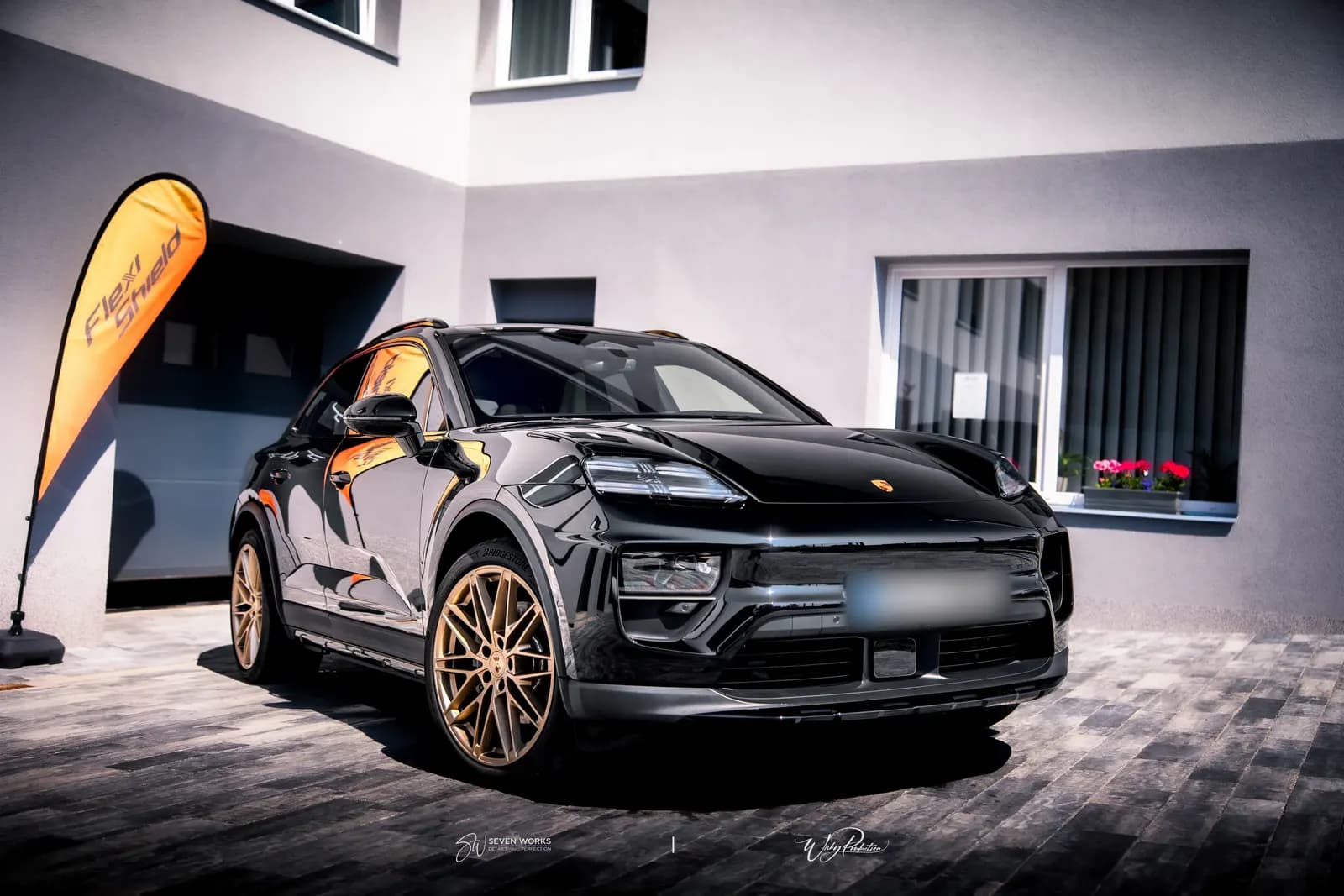 Front three-quarter view of a black Porsche SUV with gold wheels and a Flexi Shield flag in the background.