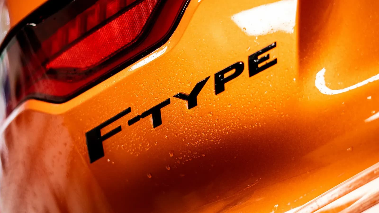 Detail zadního světla a nápisu F-TYPE na oranžovém voze, kapky vody na laku.