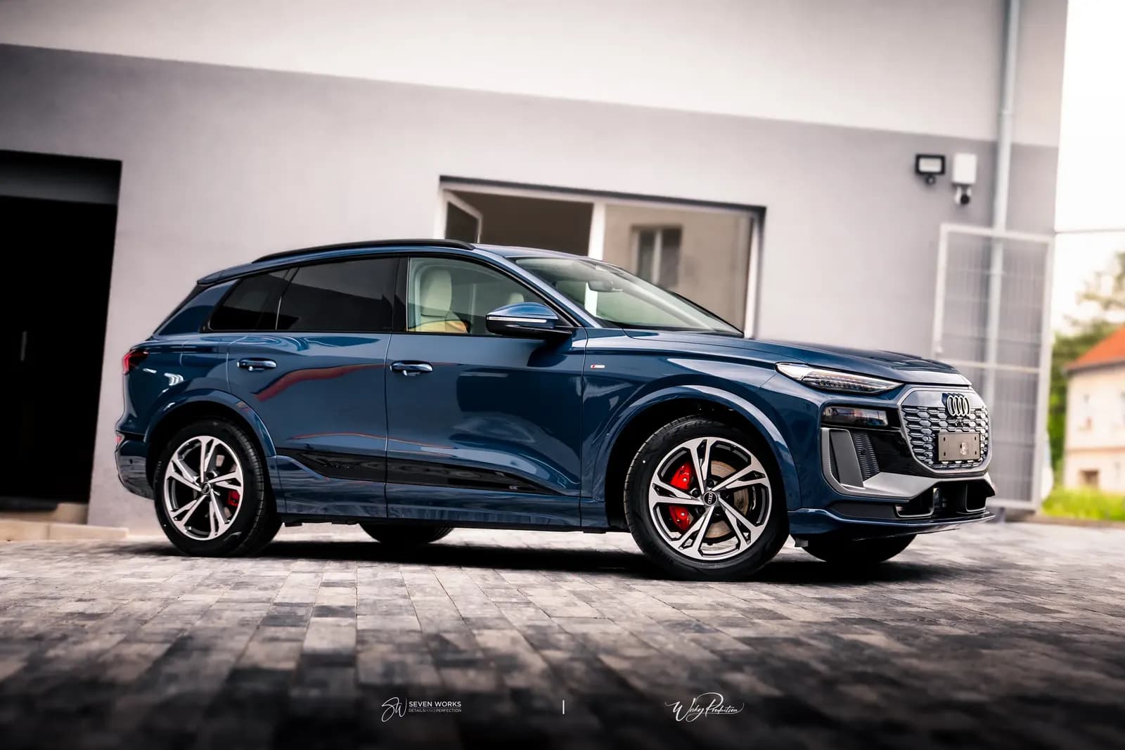 Boční pohled na tmavě modré Audi SUV s předním kolem, v pozadí dům a plot.
