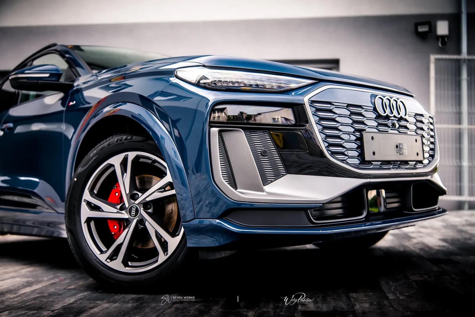 Detail předního rohu tmavě modrého Audi SUV se světlem, maskou a diskem s červeným třmenem.