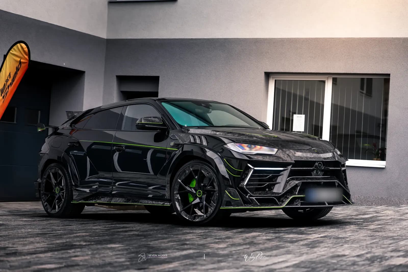 Lamborghini Urus s karbonovým widebody kitem zepředu zboku před budovou, vlevo vlajka FlexiShield.