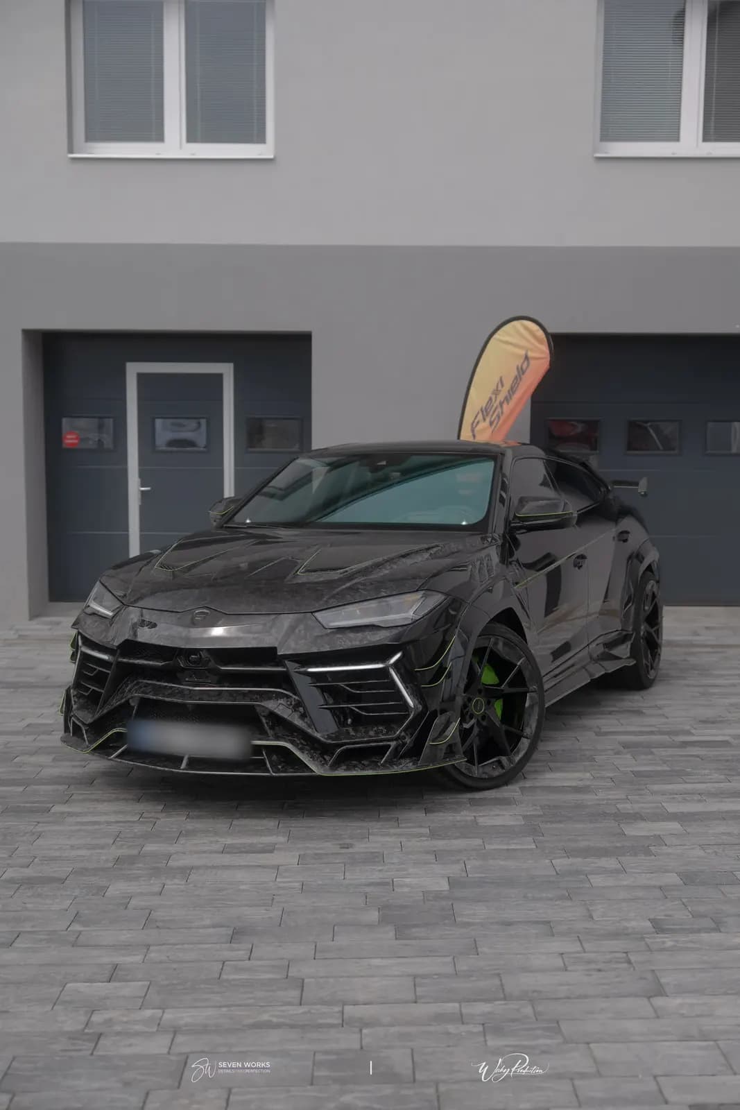 Lamborghini Urus zepředu s reklamní vlajkou FlexiShield v pozadí před dvorem s garážemi.