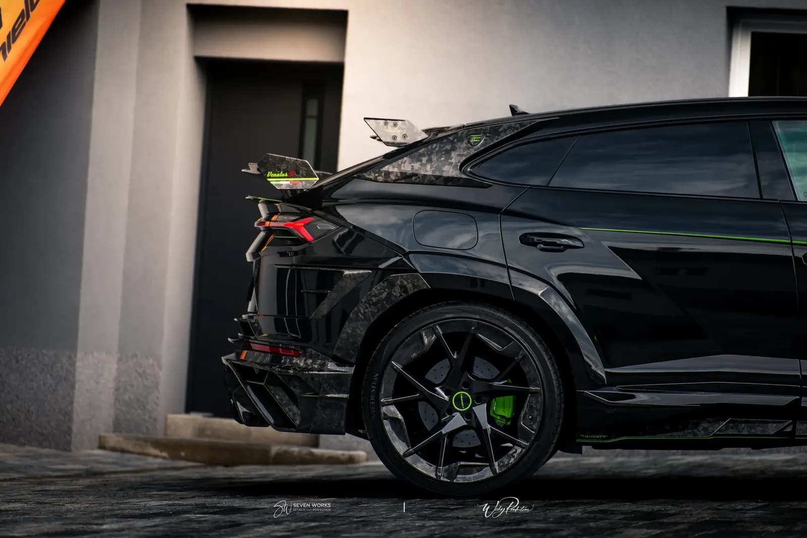 Zadní partie Lamborghini Urus se spoilerem a kolem, v pozadí vlajkový banner.