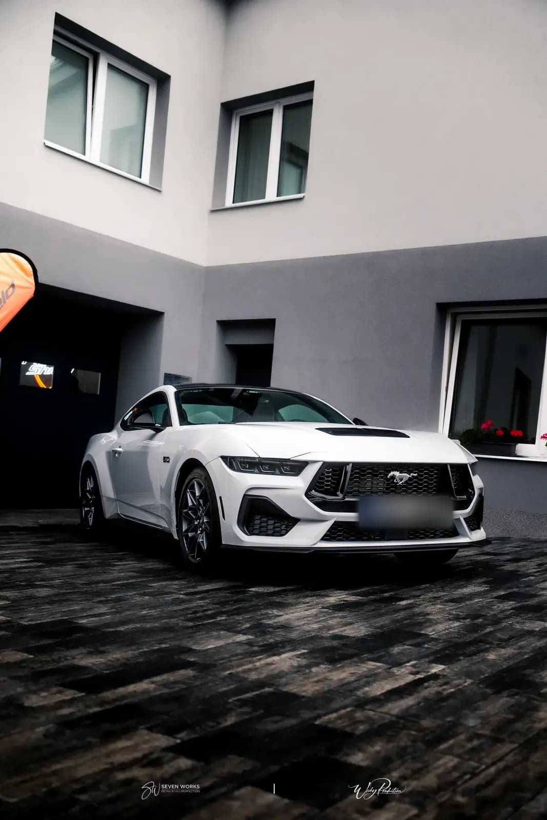 White Ford Mustang seen from the front and side on a paved courtyard, an orange lit sign behind it and red flowers in the window.