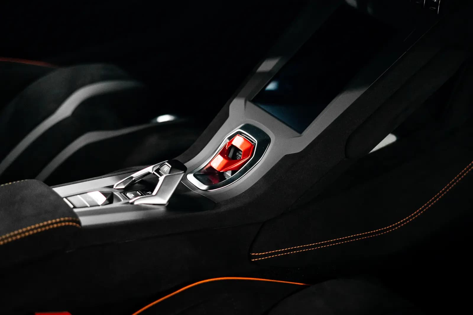 Centre tunnel of a sports car with a red lever, metal surround and orange seat stitching.