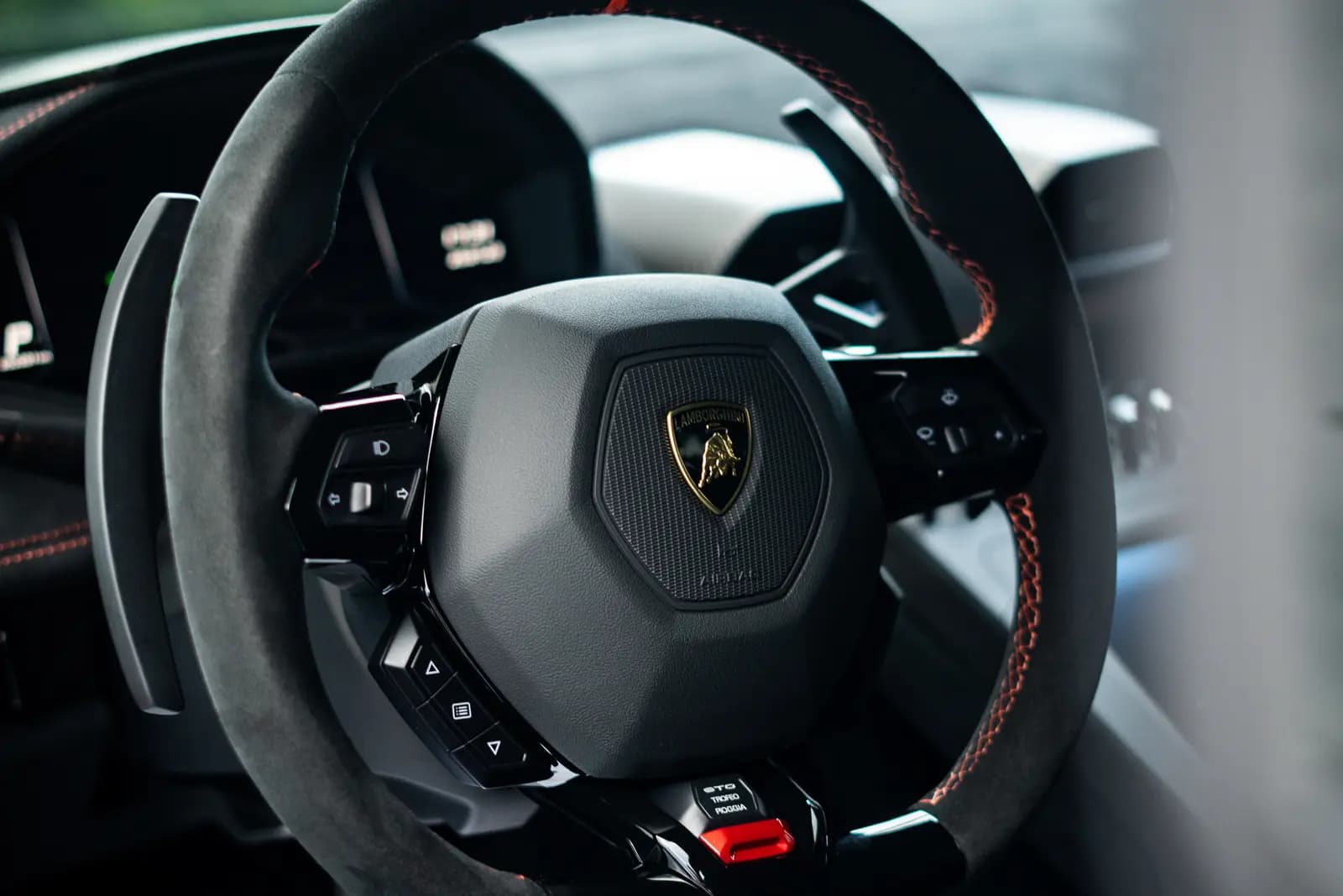 Steering wheel with the Lamborghini shield, black Alcantara and orange stitching, shift paddles below.