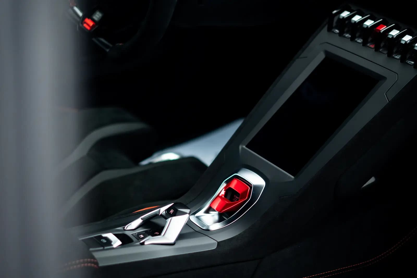 Centre console with a red switch under a chrome surround, a dark screen and orange seat stitching.