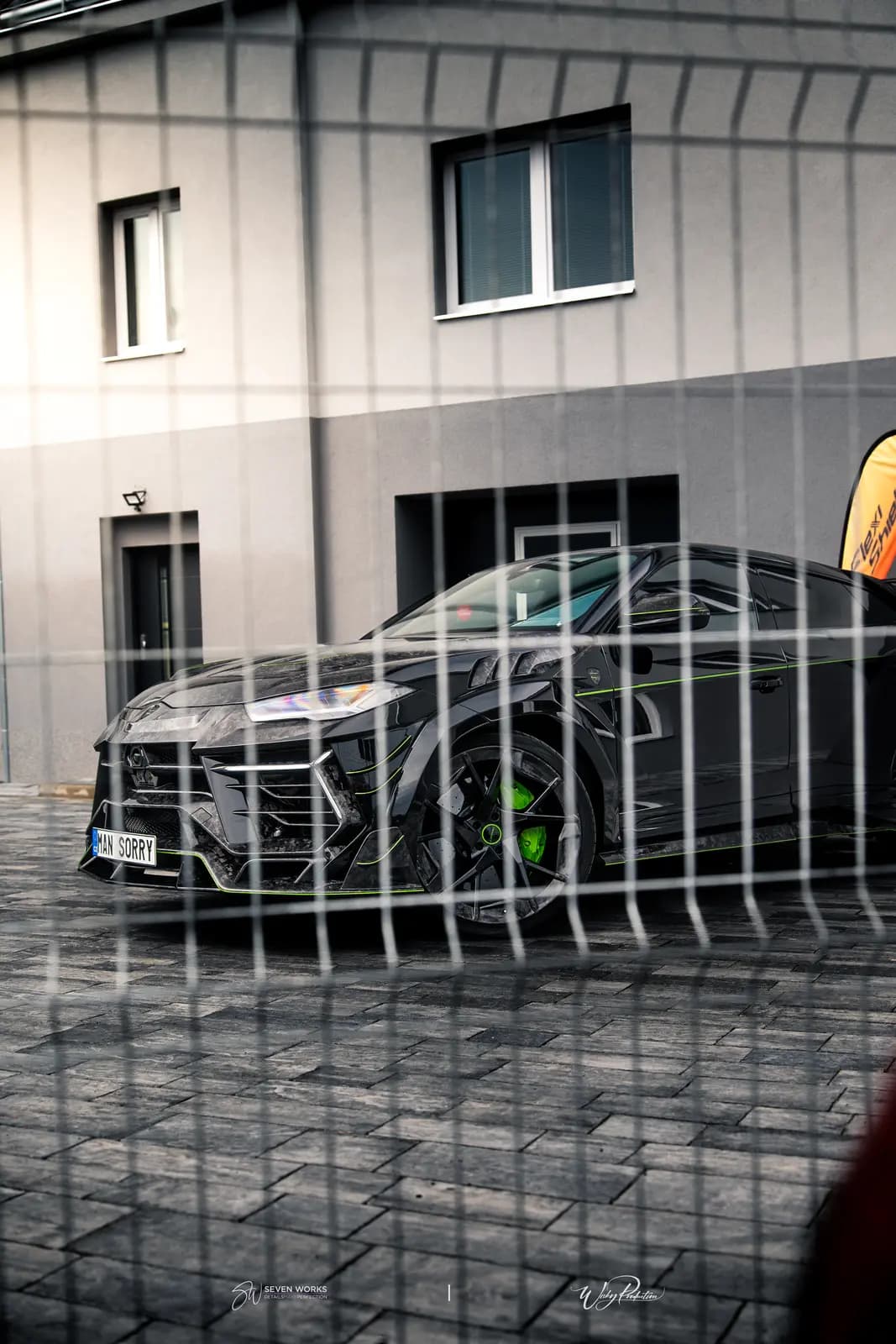 Black modified sports SUV shot through a metal fence on stone paving, a green brake caliper on the front wheel and the edge of an orange flag at right.