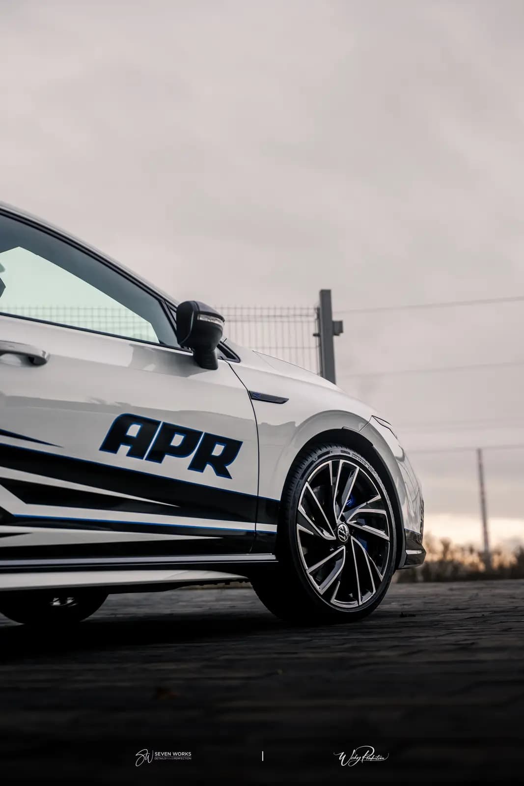 Side view of a white car with a black and blue graphic and APR lettering on the door, a blue badge on the rim, a wire fence and overcast sky behind.
