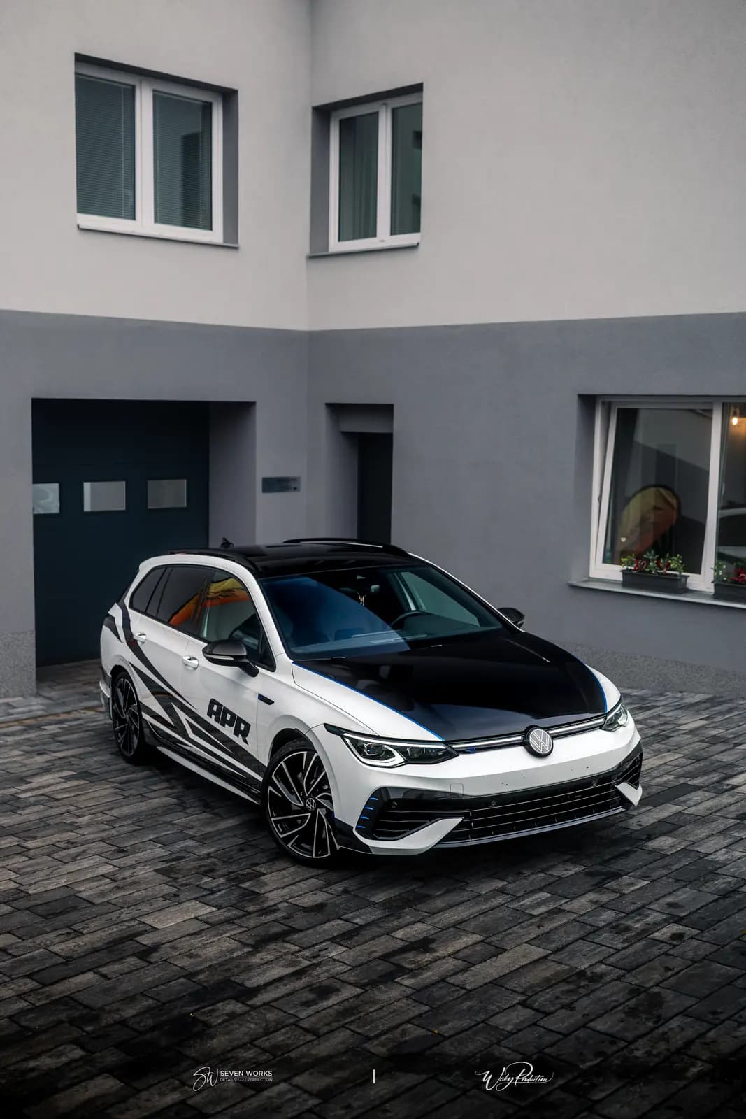 Front three-quarter view of a white car with a black bonnet and roof and APR lettering on the door, parked on paving in front of an open garage.