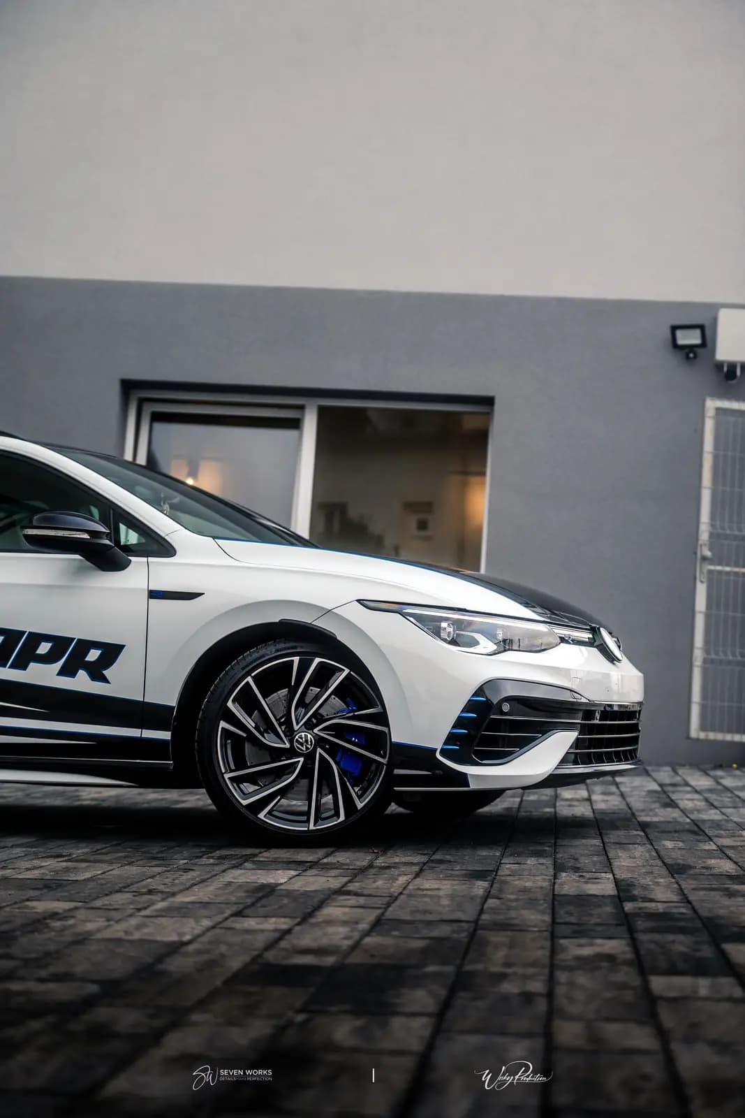Front corner with the Volkswagen badge on the grille, a blue brake caliper and part of the APR lettering on the wing, by a grey wall with windows.