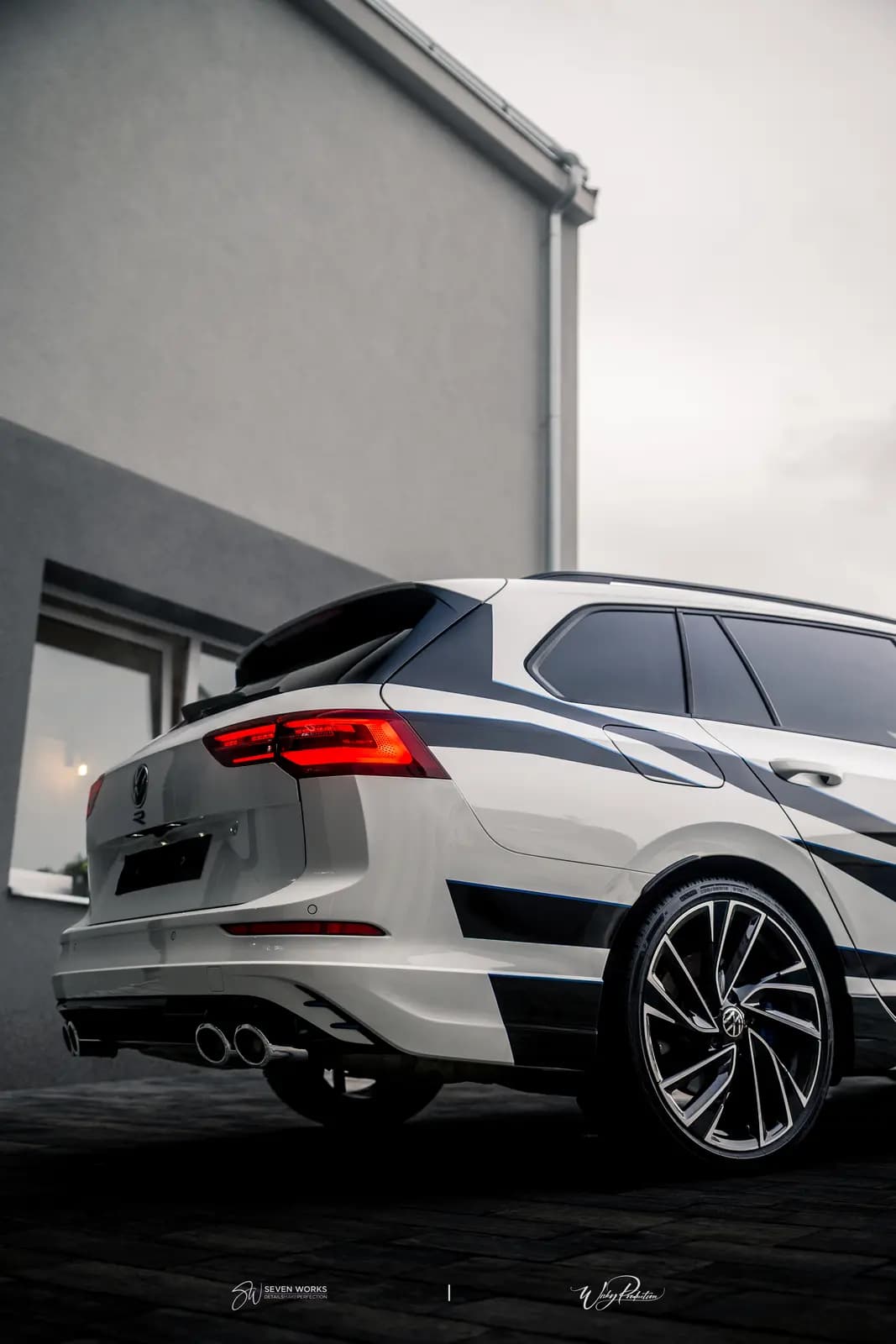 Rear view of a white car with the Volkswagen badge on the tailgate, a blue and black stripe graphic and twin exhausts, by a house under an overcast sky.