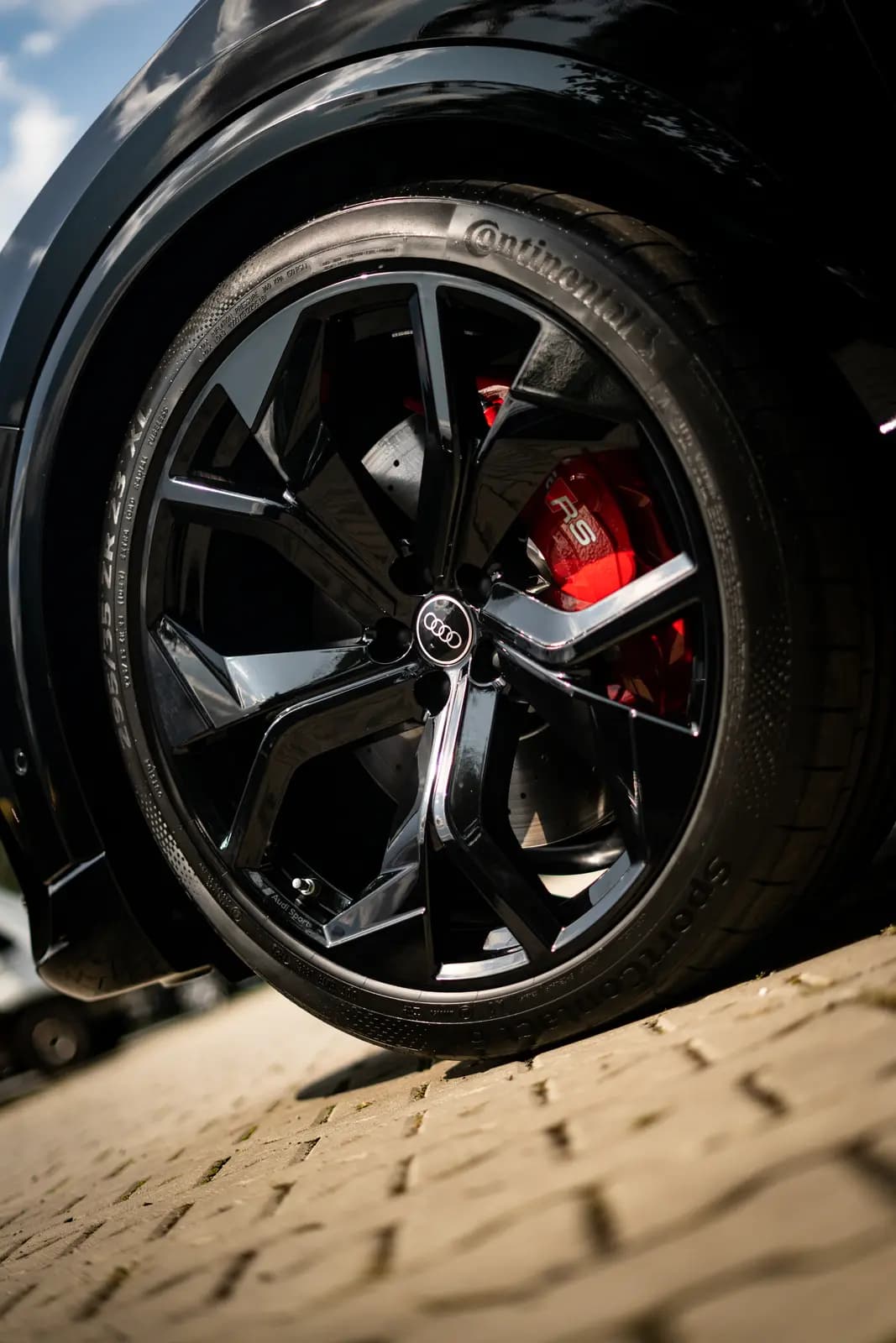 Front wheel of a black car with a red brake caliper, the Audi badge on the centre cap and a Continental tyre, outdoors on paving.