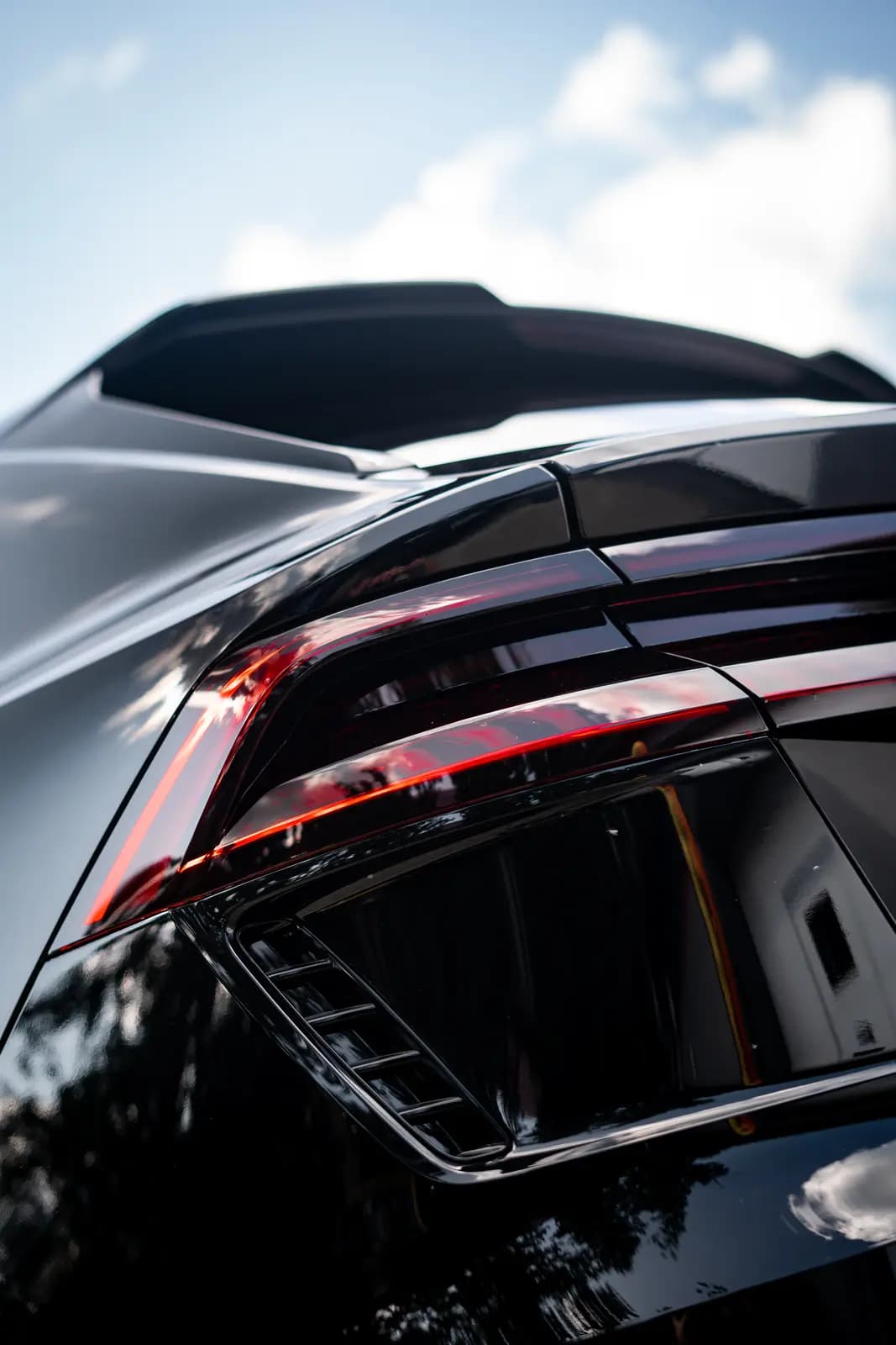 Tail light, spoiler and paintwork of a black car outdoors, sky and trees mirrored in the body.