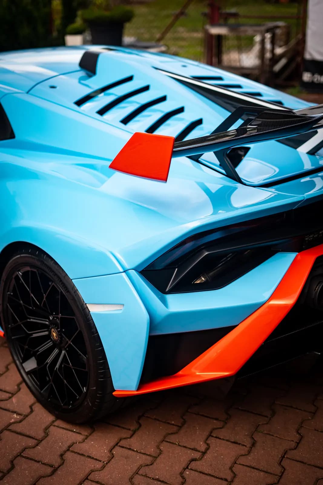 Rear of a light blue sports car with orange accents, a carbon wing and a black wheel, on brick paving with a garden behind.