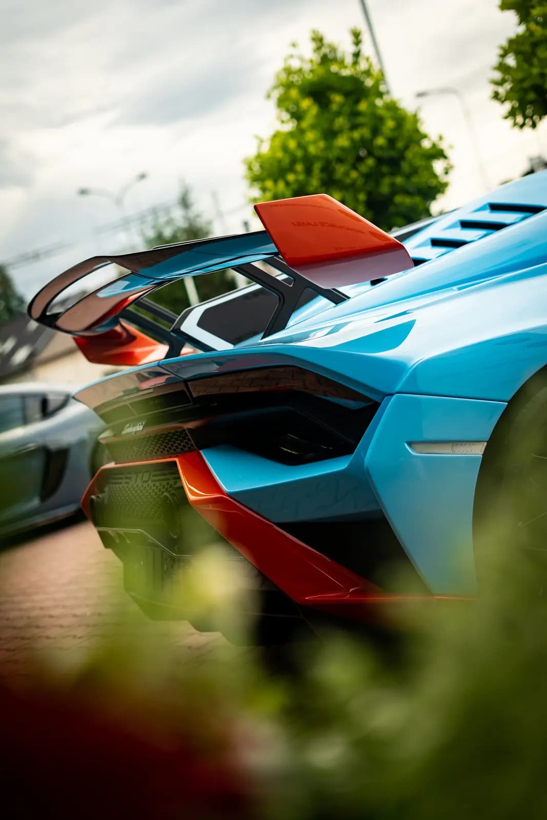 Rear of a light blue sports car with an orange spoiler and Lamborghini lettering on the boot lid, another car and trees blurred behind under an overcast sky.