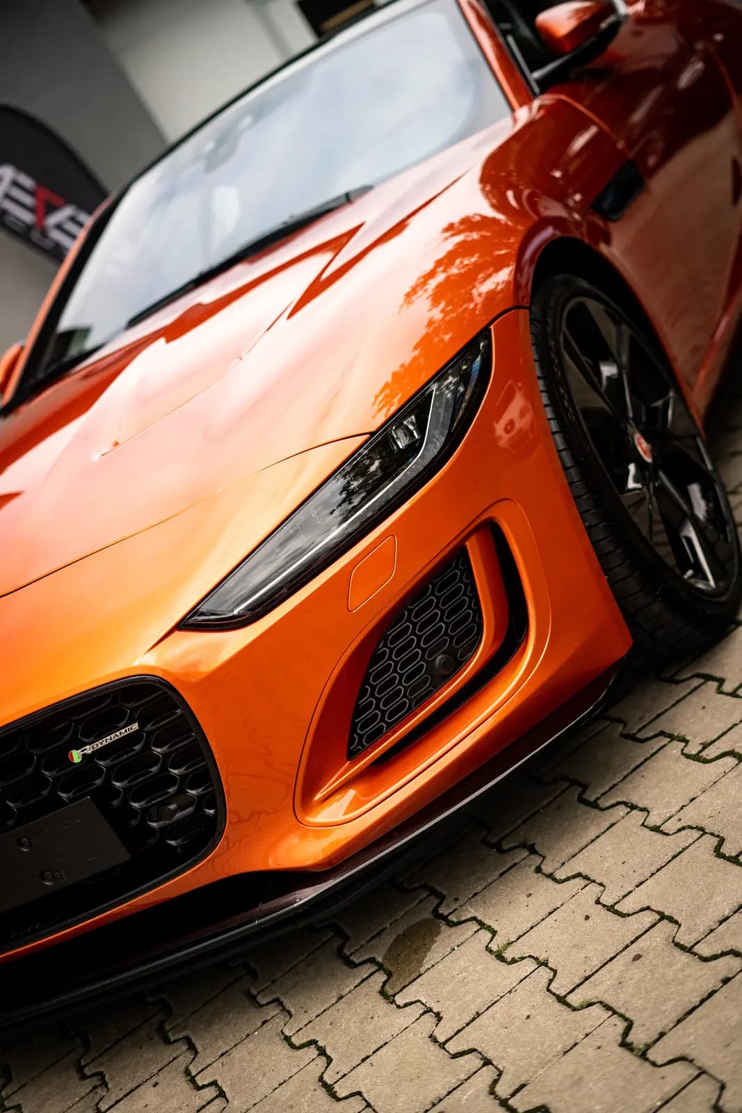 Front of an orange sports car with a black rim, parked on paving in front of a building with a blurred sign.