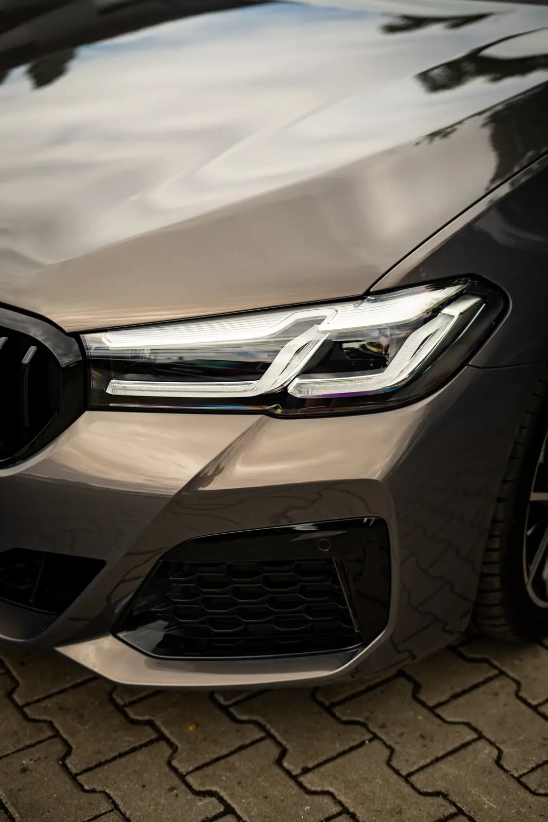 Headlight and wing of a car in grey-brown, a black wheel blurred behind, outdoors on paving.
