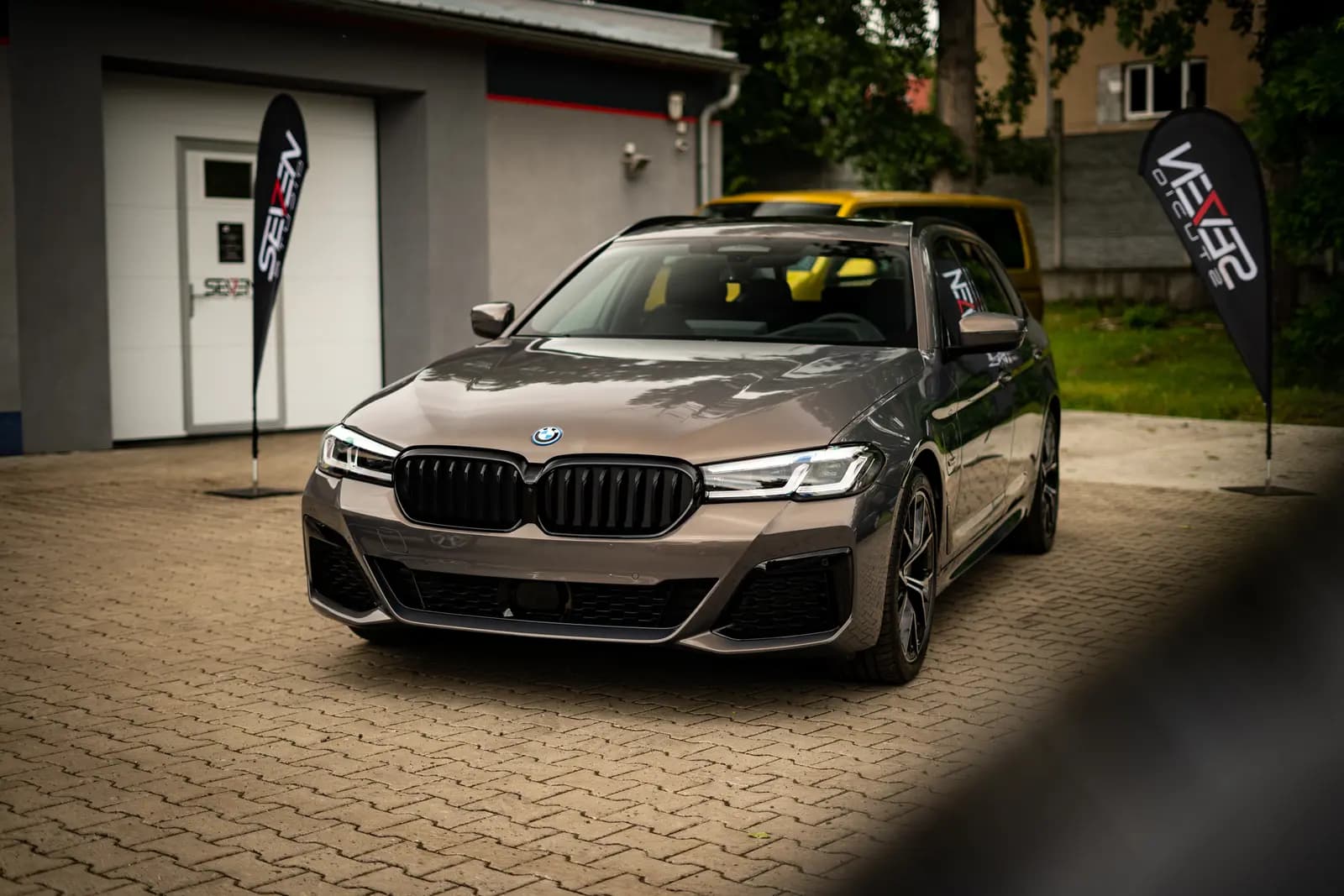 Grey-brown BMW estate seen from the front and side with the BMW badge on the grille, parked on paving in front of a building with two branded flags, a yellow car behind.