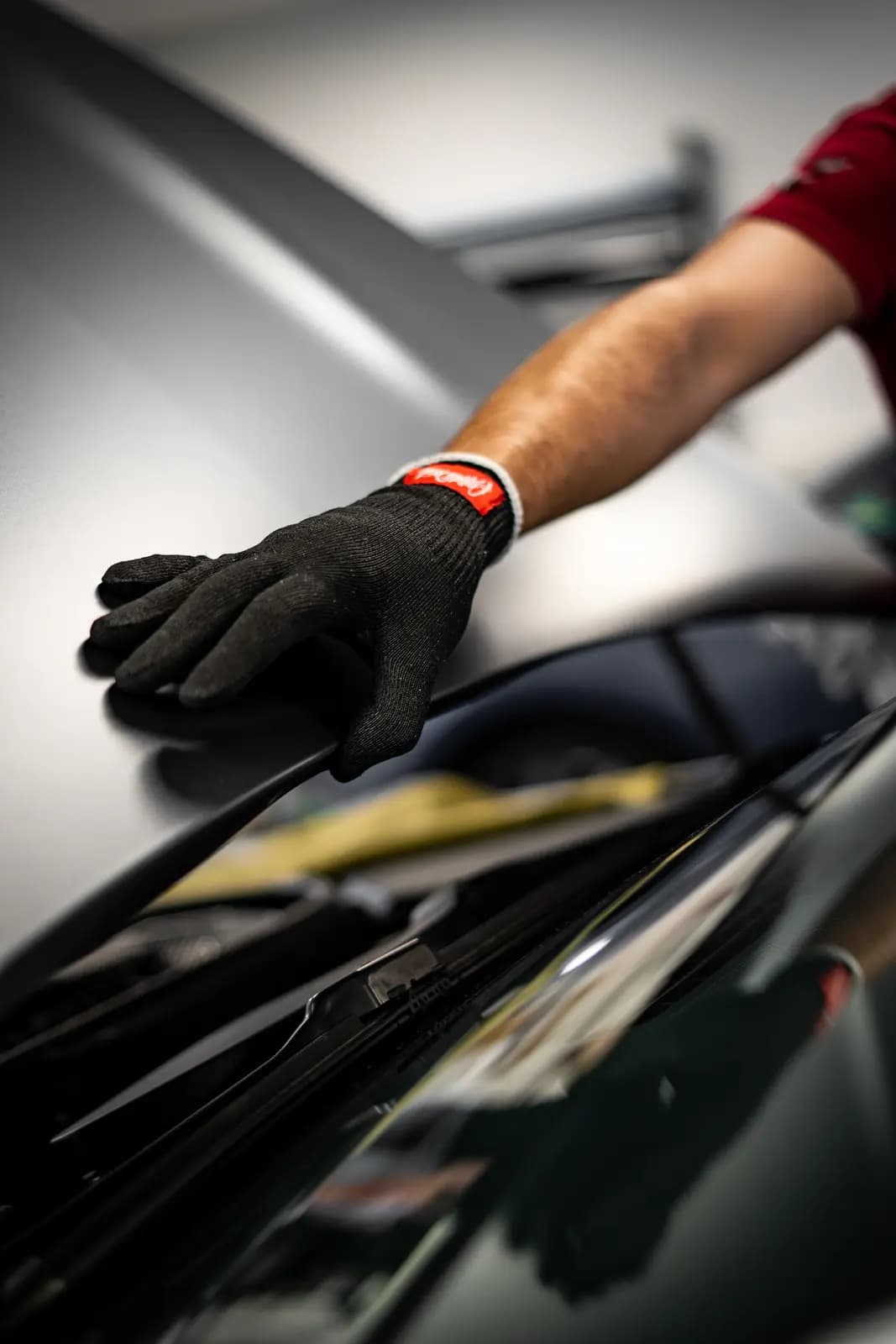 Hand in a black work glove wiping the roof of a silver car, a red sleeve on the arm, workshop equipment blurred behind.
