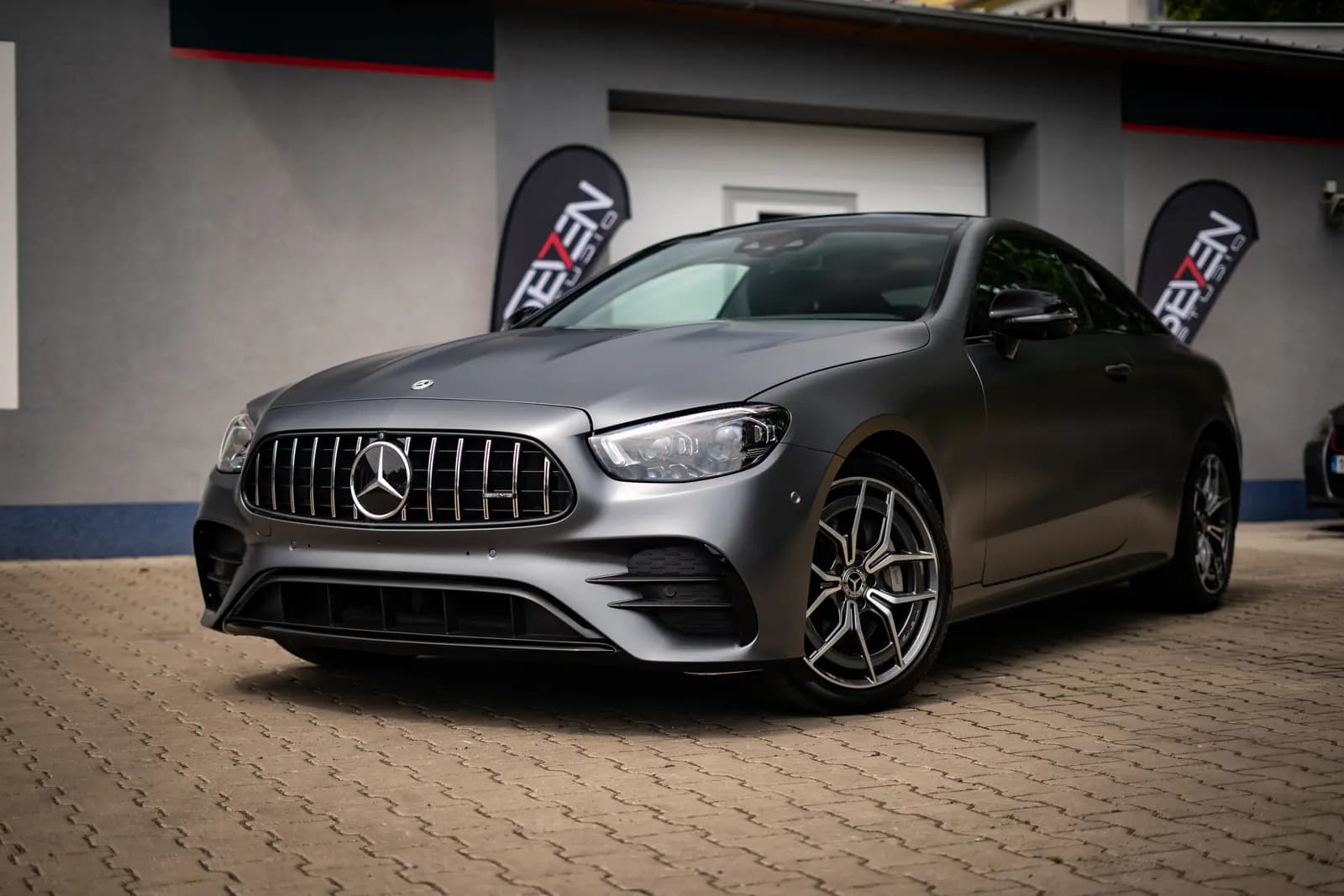 Matte grey Mercedes-AMG coupe from the front, parked on paving in front of a building with branded flags.