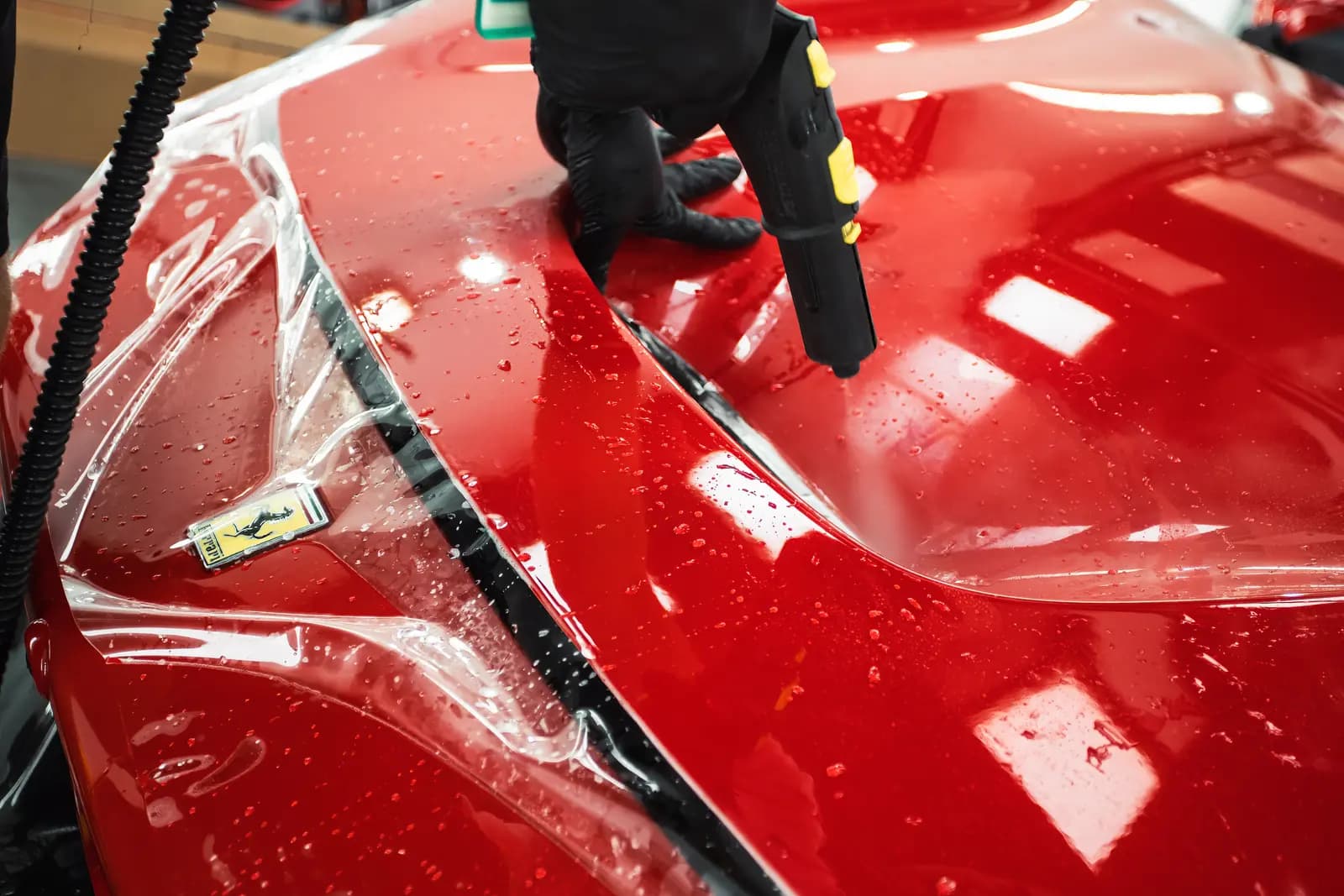 Protective film applied to the bonnet of a red Ferrari with a heat gun, the technician’s hands in black gloves.