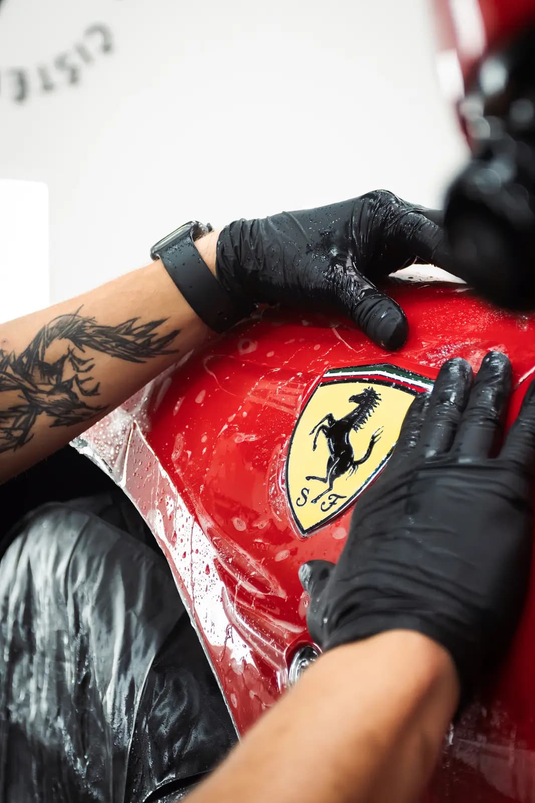 Ferrari shield badge on red bodywork, the technician’s gloved hands applying film around it.