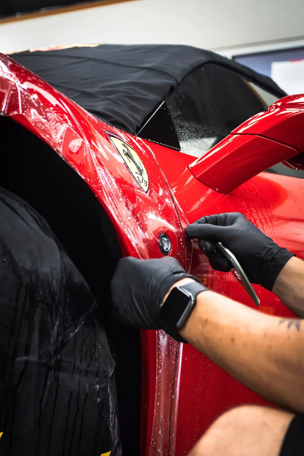 Technician squeegeeing protective film around the door and mirror of a red sports car, the body covered with a black sheet.