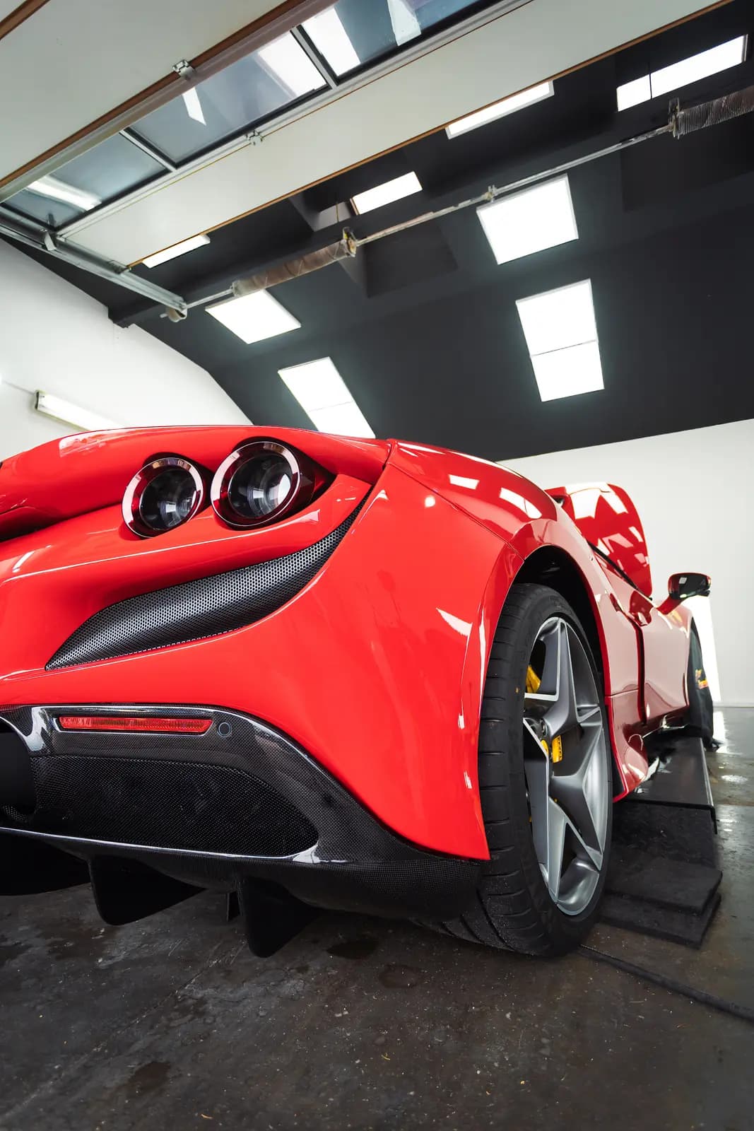 Rear of a red sports car with round lights on a ramp in a hall with roof lights, showing the wheel and diffuser.