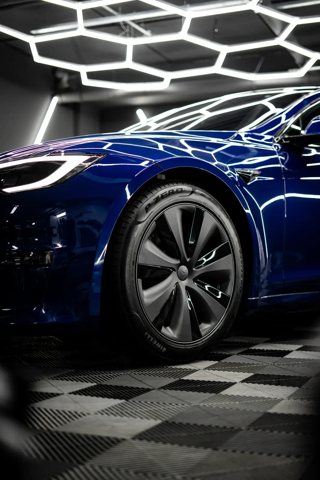 Front wheel of a dark blue car with a Pirelli P Zero tyre under honeycomb lighting.