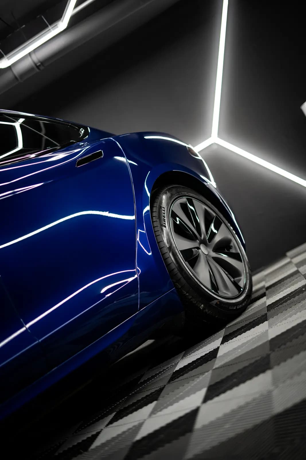 Front corner and wheel of a dark blue car, geometric light elements on the wall and a chequered floor behind.