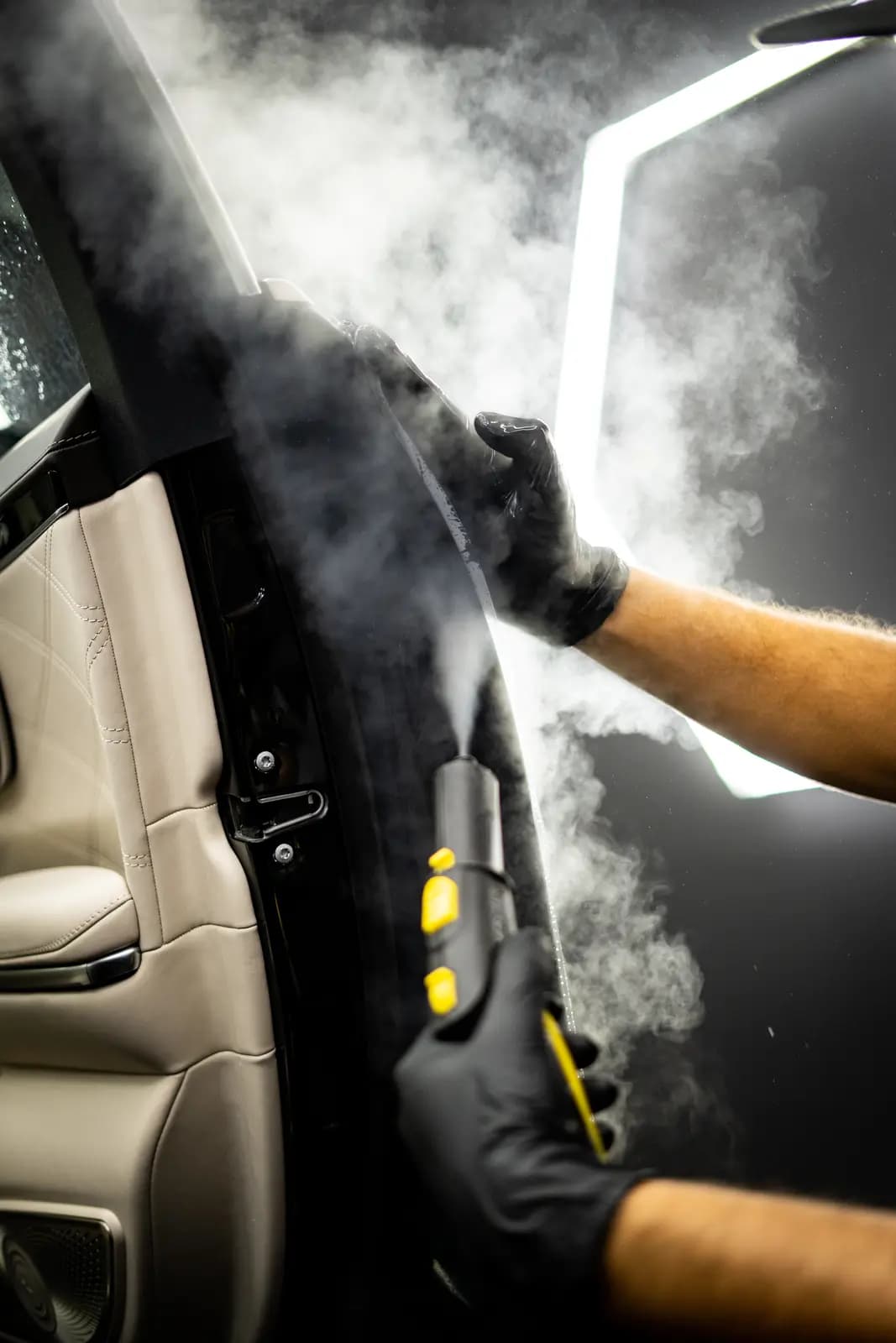 Car door seen from inside, a technician in black gloves cleaning the seal with a steam cleaner, a beige leather seat alongside.