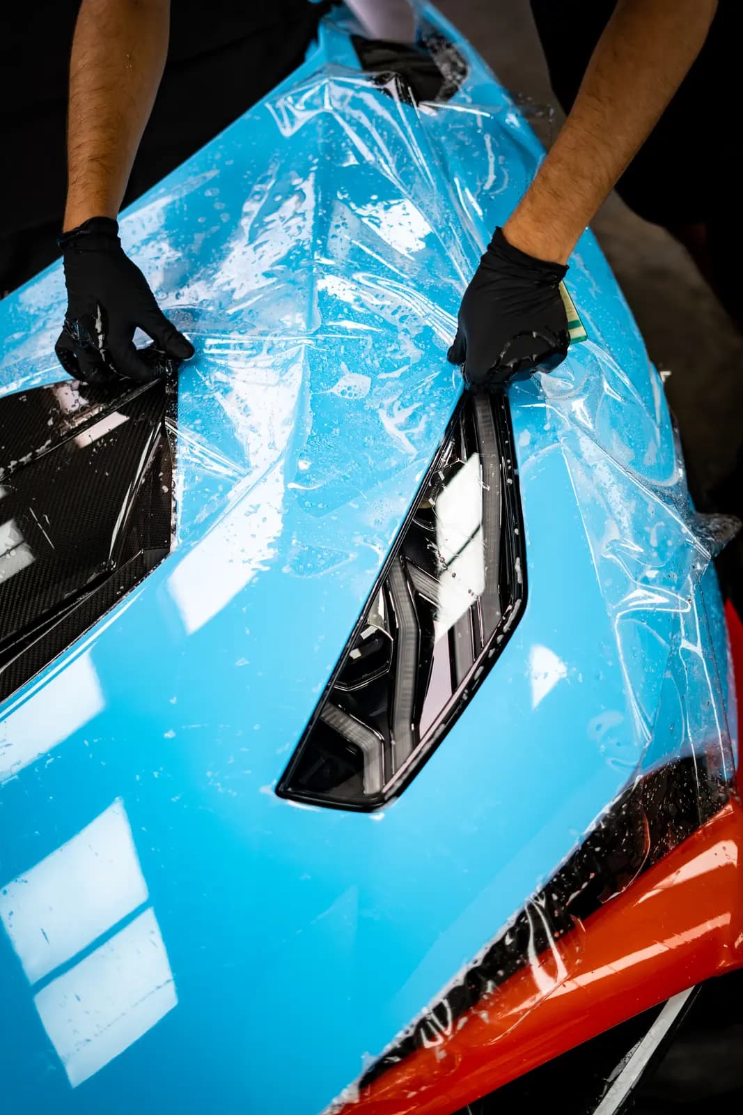 Technician in black gloves applying clear protective film to a blue car with a carbon part, a black LED headlight and an orange element below the bumper.
